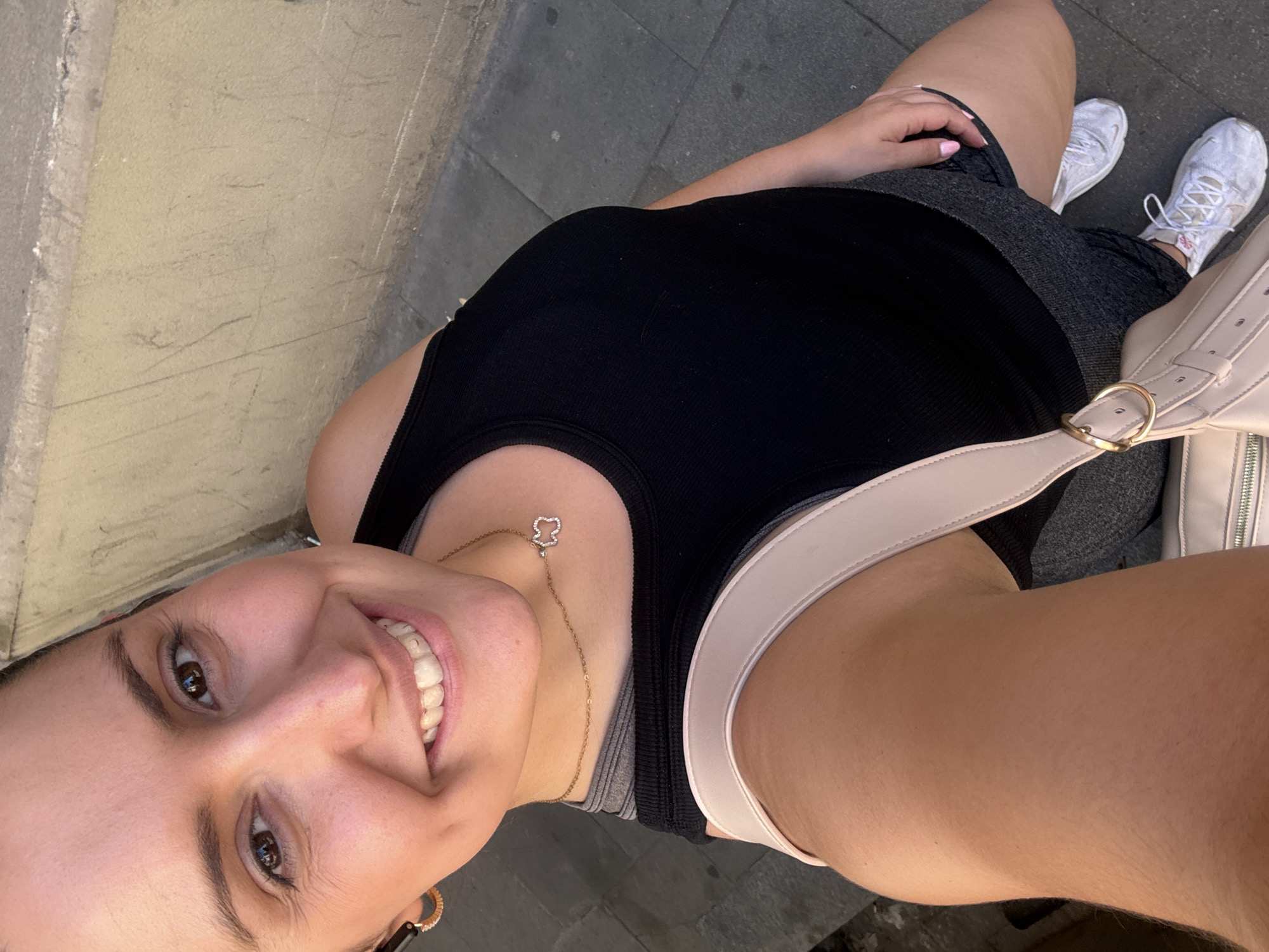 This is a selfie taken outdoors, likely during the daytime given the bright lighting. The subject is a woman with brown eyes and dark hair, smiling broadly at the camera. She is wearing a black tank top, grey athletic shorts, and white athletic shoes. A light pink crossbody bag with a gold buckle is visible, slung over her shoulder. She is also wearing a delicate gold necklace with a small charm. Her fingernails are painted a light pink color. The background shows a textured beige wall and a dark grey paved ground, consistent with an urban setting. There are faint markings on the wall. The image conveys a sense of casualness and happiness. The context provided indicates the location is Barcelona, Spain.
