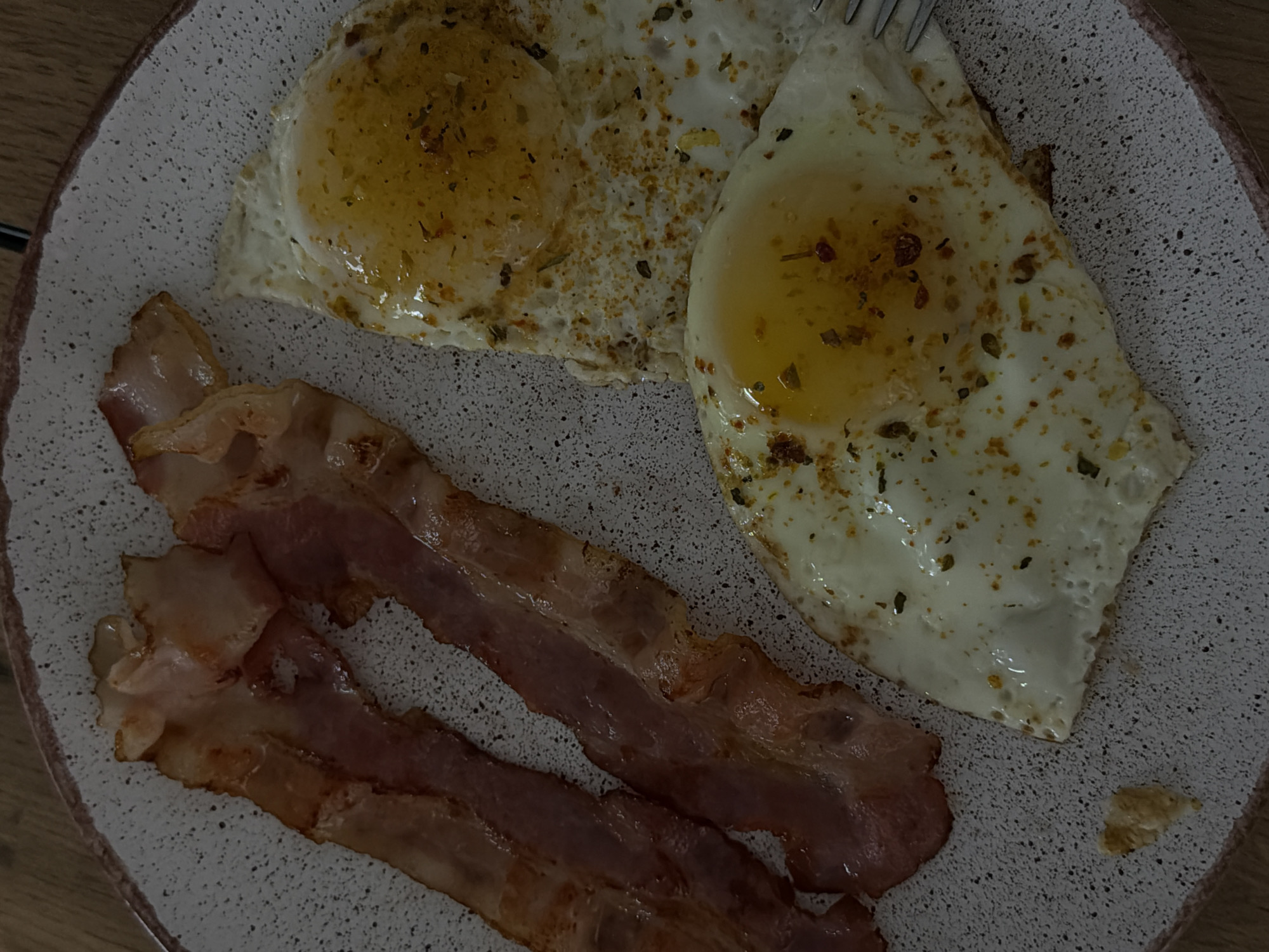 The image shows a plate of breakfast food. There are two fried eggs seasoned with herbs or spices, and three strips of bacon. The plate is speckled with gray and white, and has a dark rim. The food appears to be photographed indoors under dim lighting, possibly on a wooden table. There is no indication of the location being Reshetnikovo, Russia.