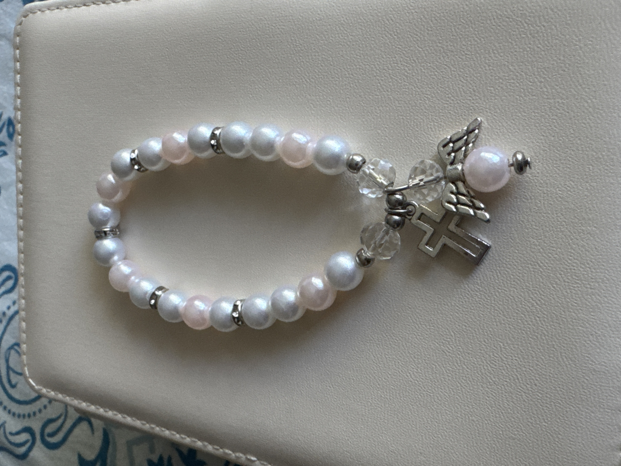 A bracelet with pearl-like beads and a silver-tone cross charm with angel wings is displayed on a textured, light-colored surface. The bracelet features alternating light pink and white beads, interspersed with small, silver-toned spacer beads. The charm is composed of a cross, two angel wings, and a small light pink bead. The background appears to be a book or notebook cover. The image does not provide any information about the city of Яромінскі сельскі Савет, Belarus, or any people.
