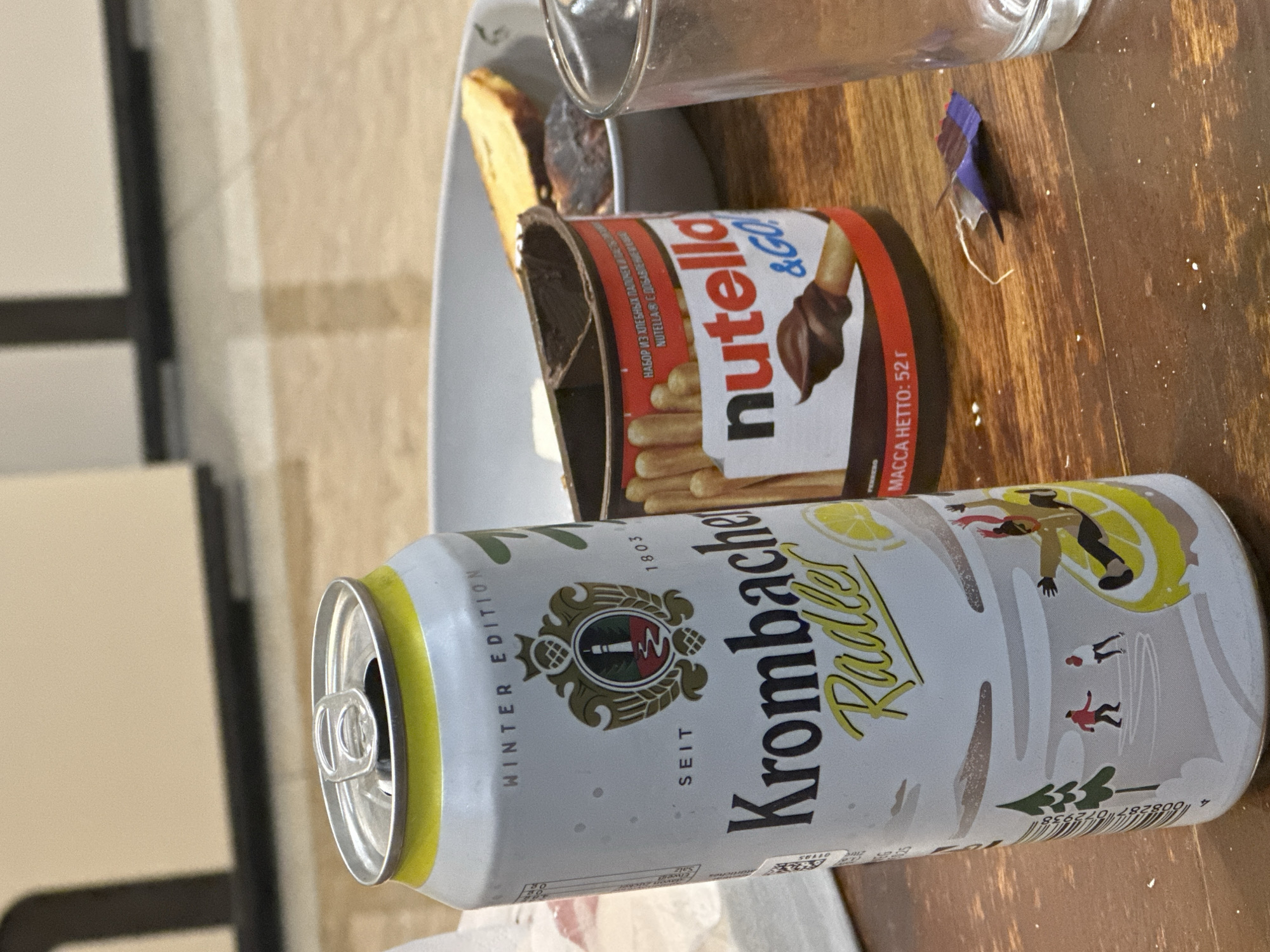 Here is a forensic description of the image:
The image shows a table surface with several items. In the foreground, there is an open can of Krombacher Radler Winter Edition beverage. Behind the can is a partially opened container of Nutella &GO! with breadsticks. To the left and behind the Nutella is a shallow plate with what appears to be a piece of food on it. Next to the Nutella is a partially full clear glass. A small, torn piece of colorful paper is present on the table surface. The table surface is wood-grained. The background is blurred, but it includes a wall and a window frame.