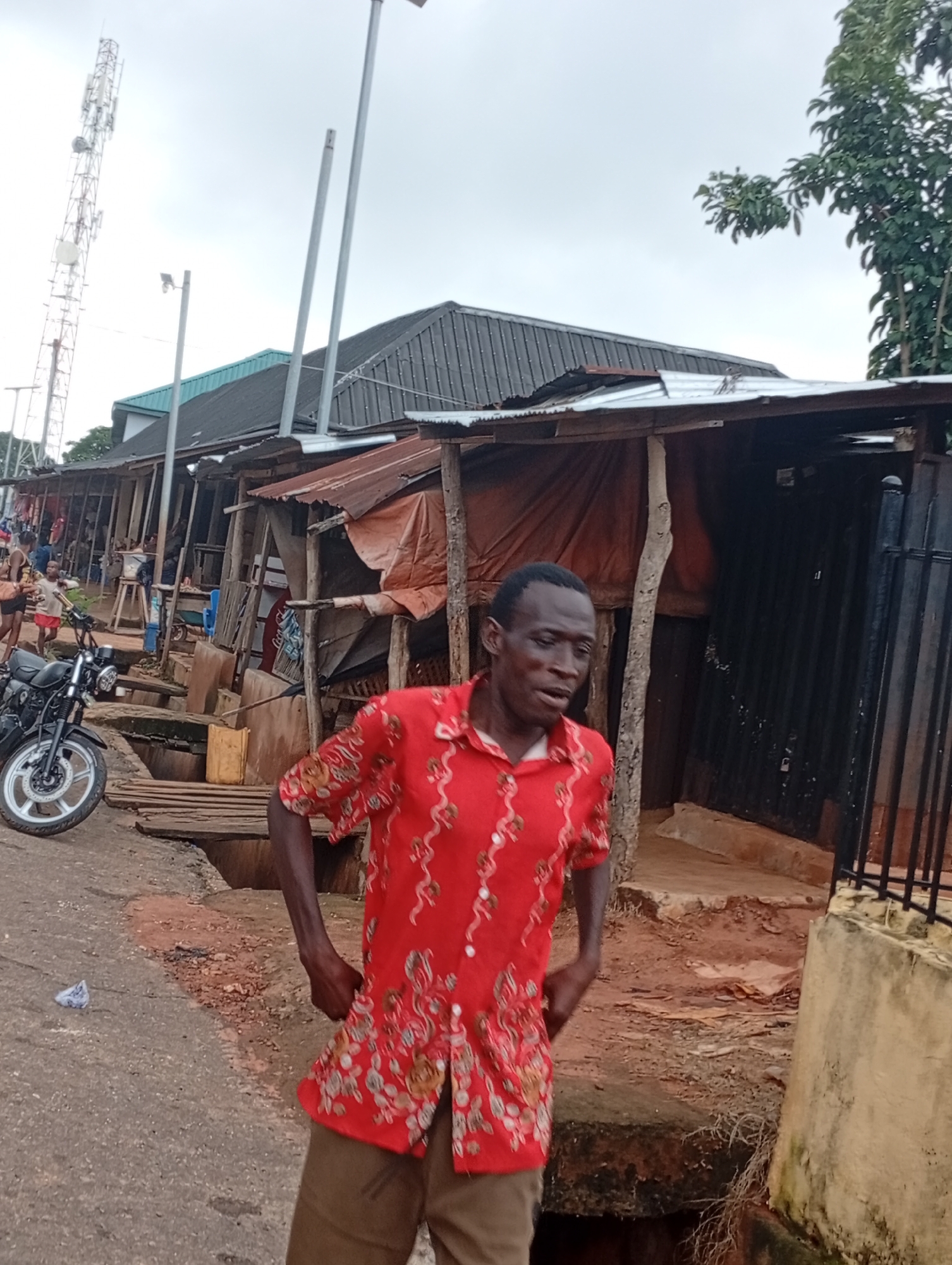 A man in a red, floral-patterned short-sleeved shirt and tan pants stands on a dirt and asphalt road. He is looking towards his right, with his mouth slightly open. His left arm is bent at the elbow, and his right arm is down by his side, with his hand in a fist. In the background, there is a marketplace with wooden stalls covered with corrugated metal and tarps. A motorcycle is parked on the left side of the road, and a cell tower is visible in the distance. The sky is overcast.