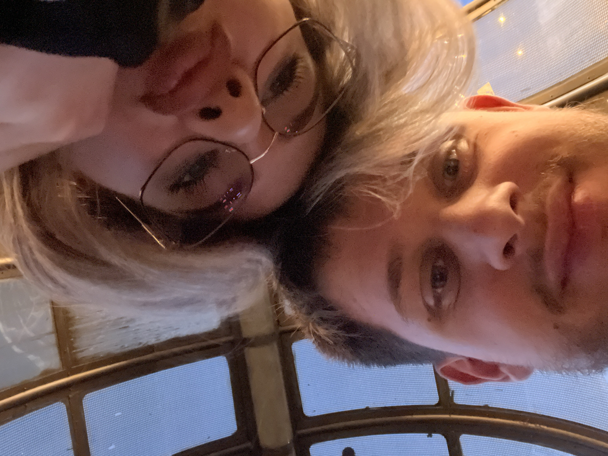 Two people, a man and a woman, are posing for a selfie indoors. The woman is on the left side of the image, and she wears glasses. The man is on the right side of the image and has short brown hair. The picture is taken from a low angle, looking up at the subjects. They are indoors, with a lattice-like structure in the background. The lattice background could possibly be a window or roof. There is no immediately evident information in the image to place the location in Blagoevgrad, Bulgaria.