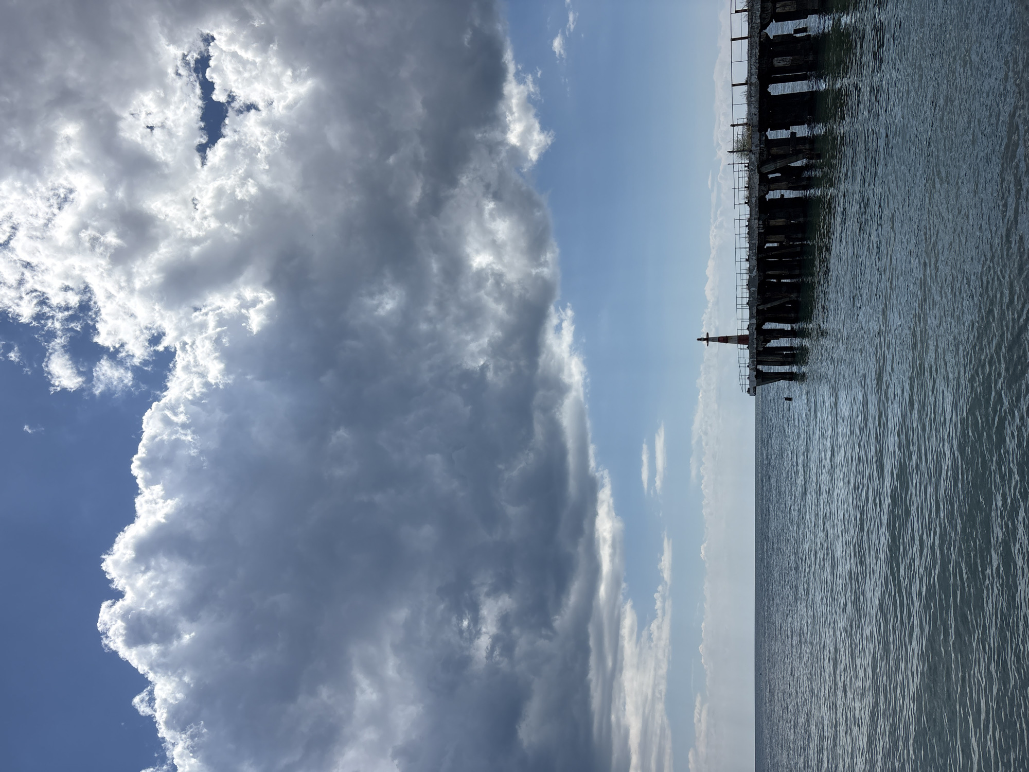 An image of a pier extending into the sea under a dramatic sky. The sky is filled with large, dark grey clouds that create a sense of foreboding, contrasted with patches of bright blue sky and white, illuminated clouds. The sea is a deep blue-green, with small waves creating ripples on the surface. The pier is made of concrete and wood, with weathered pillars supporting the structure. A small red and white lighthouse stands at the end of the pier. The image appears to be taken from a low angle, looking up towards the sky and out towards the sea. The scene is serene and empty, with no people or animals visible. The image is likely taken in Alushta, Ukraine, a coastal city known for its scenic beauty and piers.
