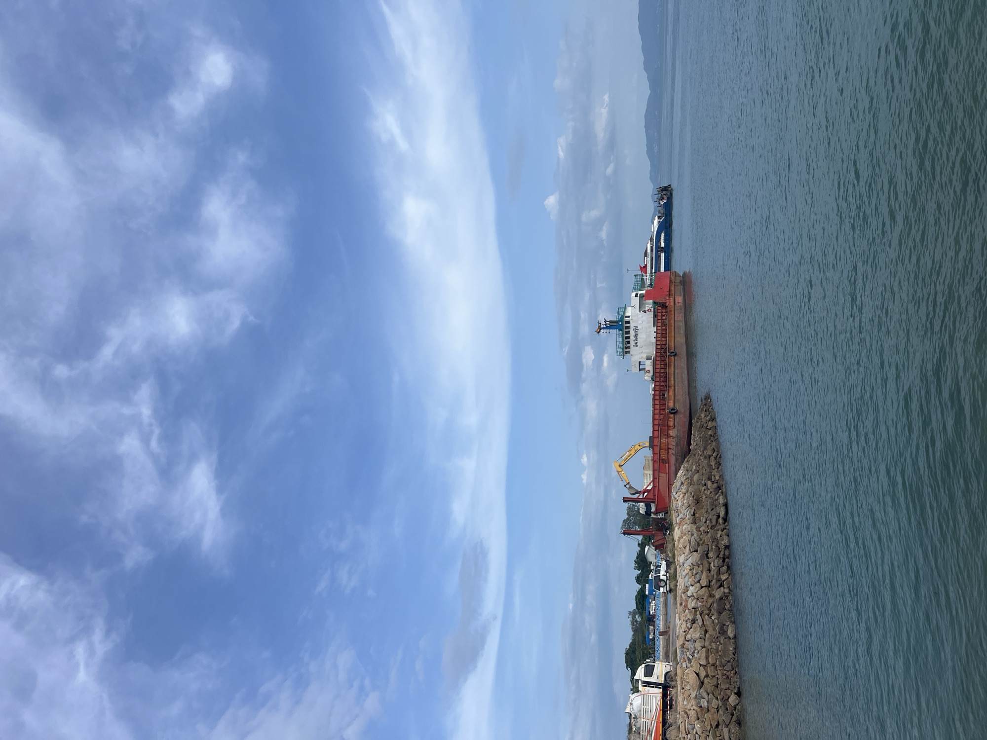The image depicts a harbor scene on the island of Ko Pha-ngan, Thailand. In the foreground, the calm, blue-green water of the sea is visible, with gentle ripples. A rock embankment lines the left side of the scene, separating the water from the land.
Moored alongside the embankment are several vessels. The most prominent is a large, red barge, partially loaded with materials and equipped with a yellow excavator. To the right of the barge, a smaller, blue and white passenger ferry is docked, with several people visible on its deck. In the distance, more boats and landmasses with green vegetation can be seen under a vast sky filled with scattered white clouds. On the shore behind the barge, there are trucks and some structures suggesting port or construction activity. The overall atmosphere is one of a working harbor on a bright, partly cloudy day.