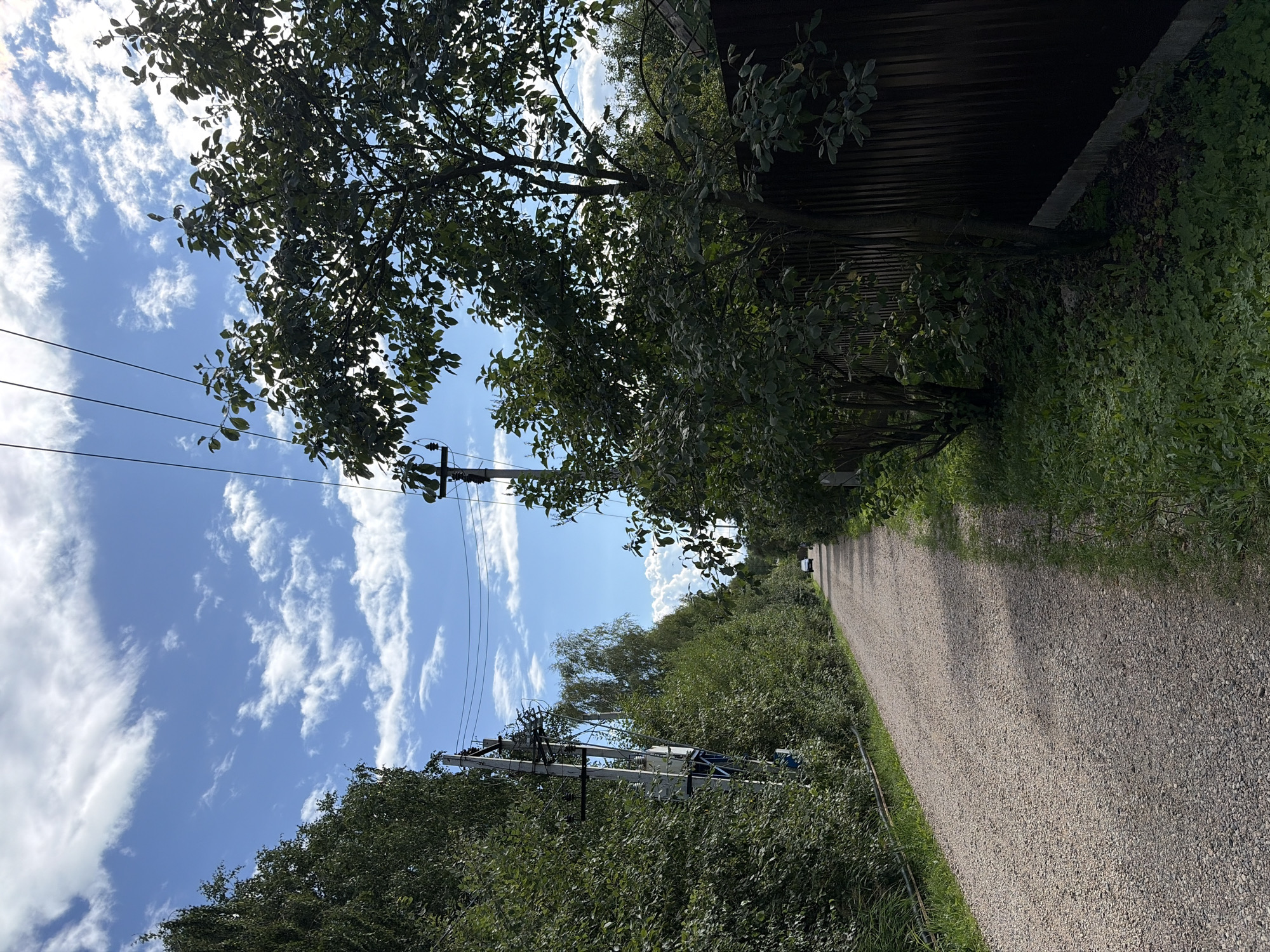 The image depicts a gravel road lined with trees and vegetation in Шахтер Снт, Russia. A white car is visible driving away from the camera in the distance. Overhead power lines and poles are present along the left side of the road. A dark brown wooden fence runs along the right side of the road, partially obscured by trees and foliage. The sky is blue with scattered white clouds, suggesting daytime. The environment appears to be a rural or semi-rural area.