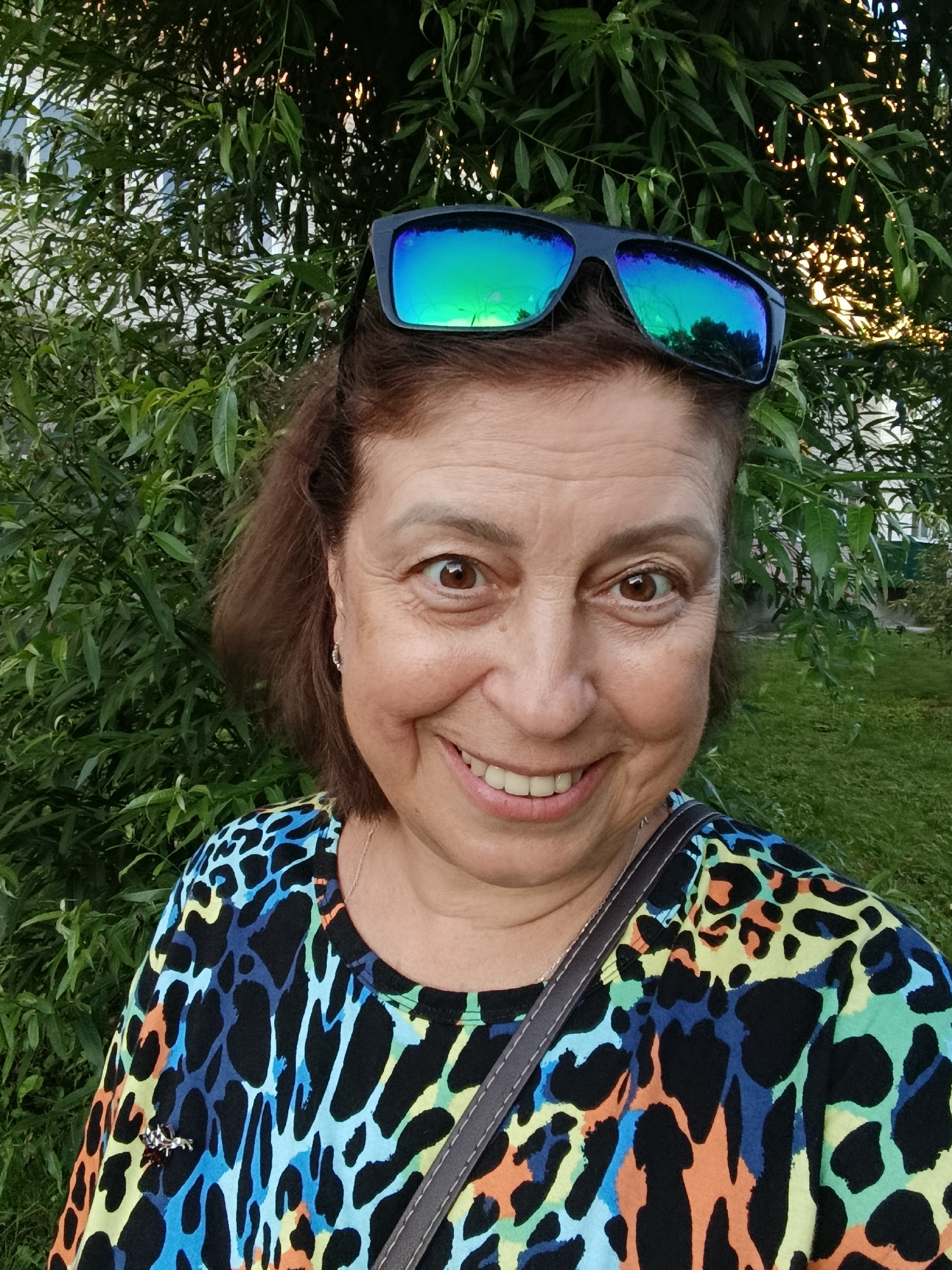 A middle-aged woman with brown hair and brown eyes smiles at the camera, wearing sunglasses perched on her head and a colorful, leopard-print shirt. She is outdoors, with lush green foliage behind her and a hint of grass and a building in the background. The lighting suggests it is daytime, possibly late afternoon, given the warm glow on the leaves. The image appears to have been taken in Vareya, Russia.