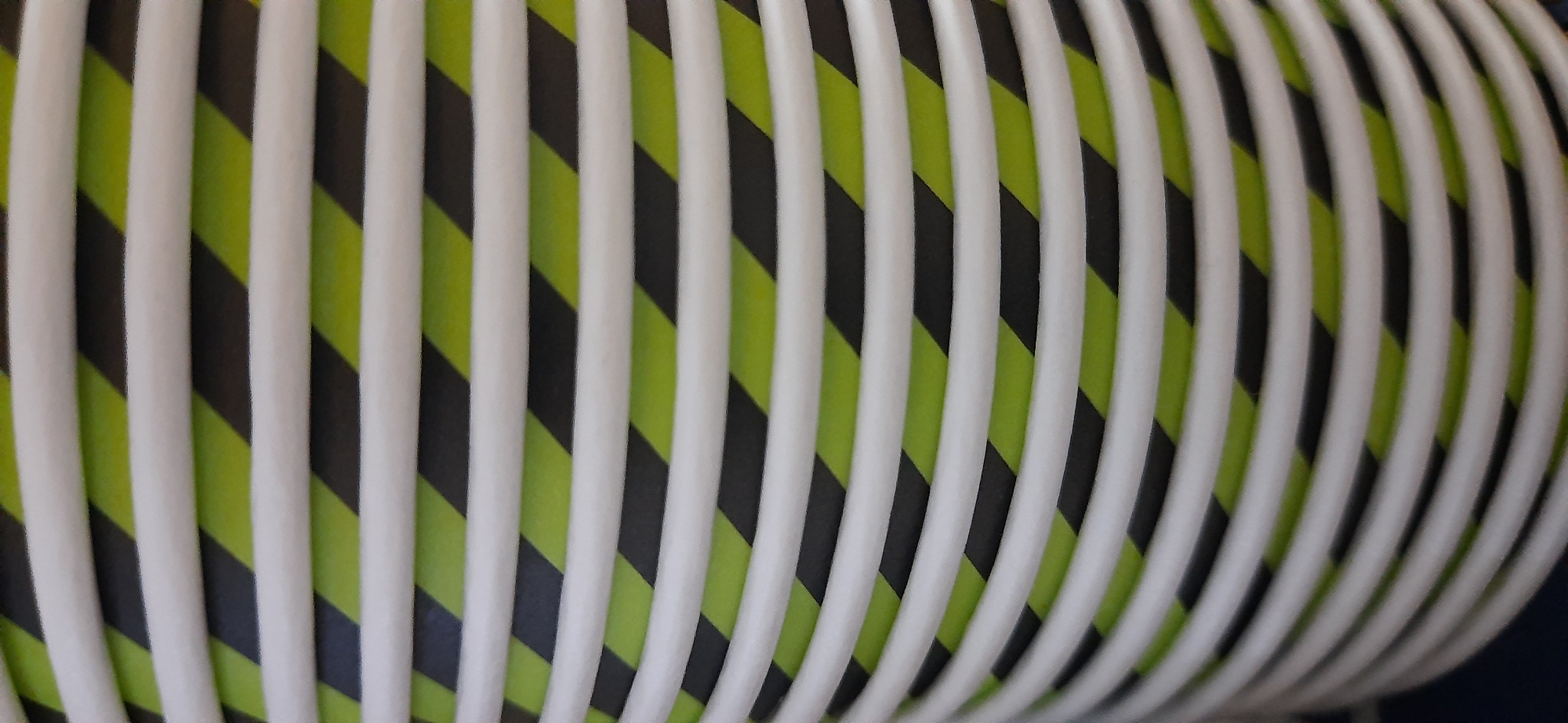 The image shows a close-up, abstract view of a stack of disposable paper cups with a black and lime green diagonal stripe pattern. The cups are stacked vertically and appear to be a uniform size and design. The lighting is soft and diffuse, highlighting the texture and slight curvature of the cups. The background is dark and out of focus, suggesting the cups are in a dimly lit environment. The image does not contain any identifiable landmarks or elements that would allow for its location to be determined in the city of Dnipro, Ukraine, or any other location.