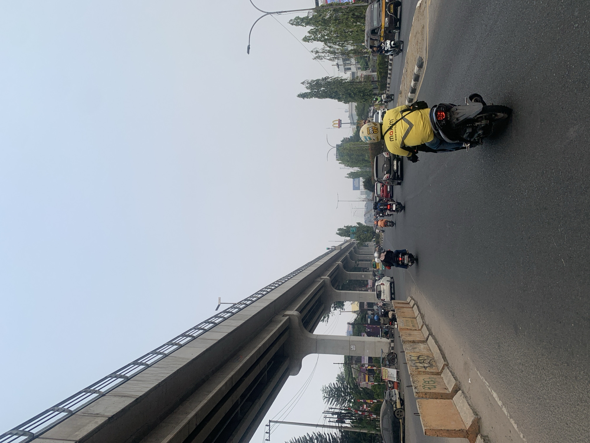 The image shows a busy street in Palembang, Indonesia, with an elevated road structure dominating the left side of the frame. A person wearing a bright yellow jacket with "maxim Rides and Delivery" written on it is riding a motorcycle in the foreground, facing away from the camera. Several other motorcycles and cars are visible on the road, indicating moderate traffic. The environment appears to be urban, with buildings and trees lining the street in the background. The sky is overcast, suggesting a cloudy day. The elevated road structure is supported by large concrete pillars and has a railing along its edge. Concrete barriers with graffiti are present along the side of the road. A McDonald's sign is visible in the distance.
