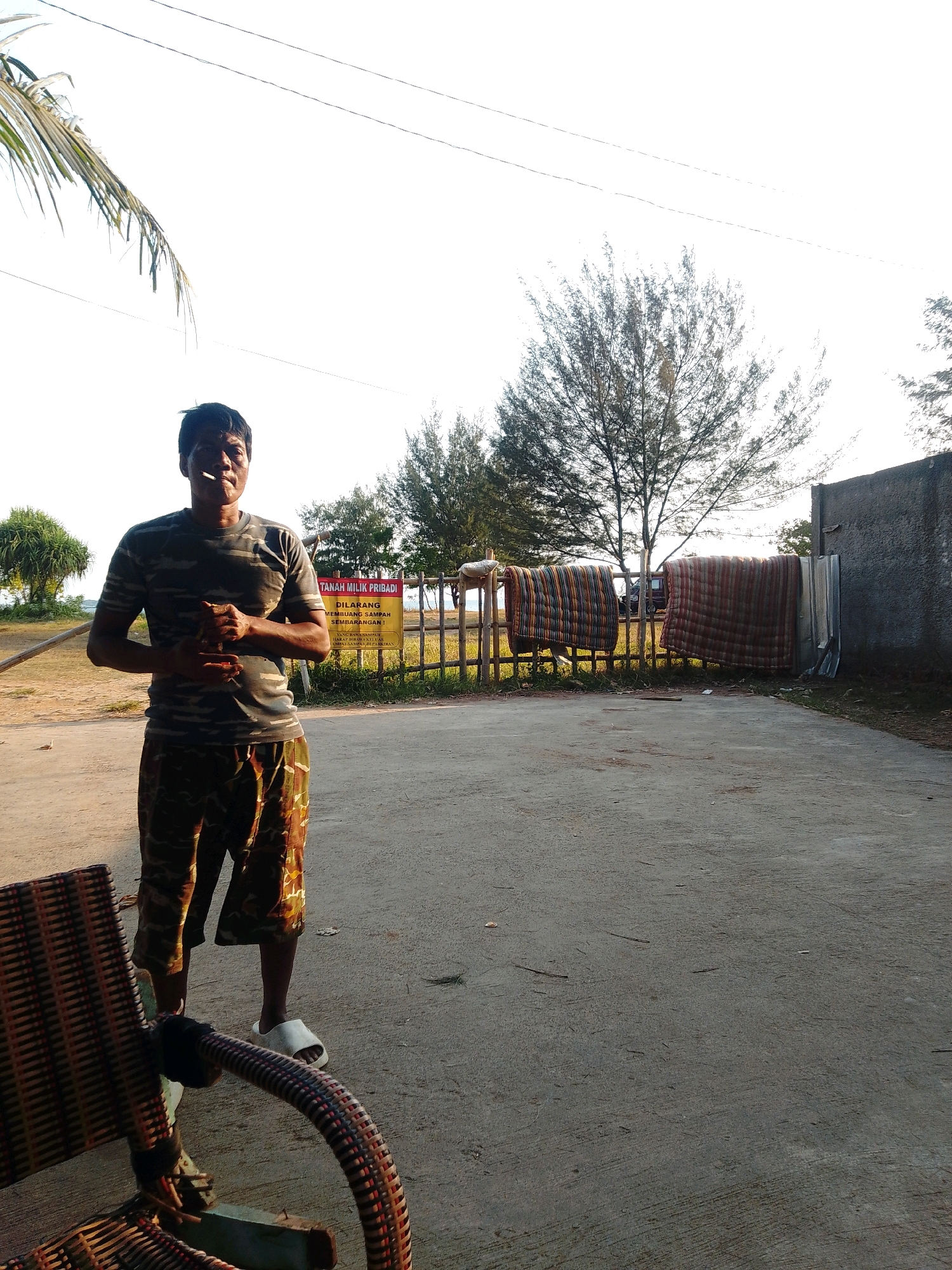 The image depicts an outdoor scene with a man standing in the foreground, holding a cigarette. He is wearing a camouflage t-shirt and camouflage shorts, and white flip-flops. Behind him, a wooden fence with a yellow sign is visible. The sign has Indonesian text that translates to "PRIVATE LAND" and "DO NOT LITTER". Several blankets or mats are hung on the fence. In the background, there are trees, a concrete wall, and power lines against a bright sky. The ground is paved with concrete. The scene appears to be in a rural or coastal area, possibly near the sea given the vegetation. The location is identified as Cikal, Indonesia.