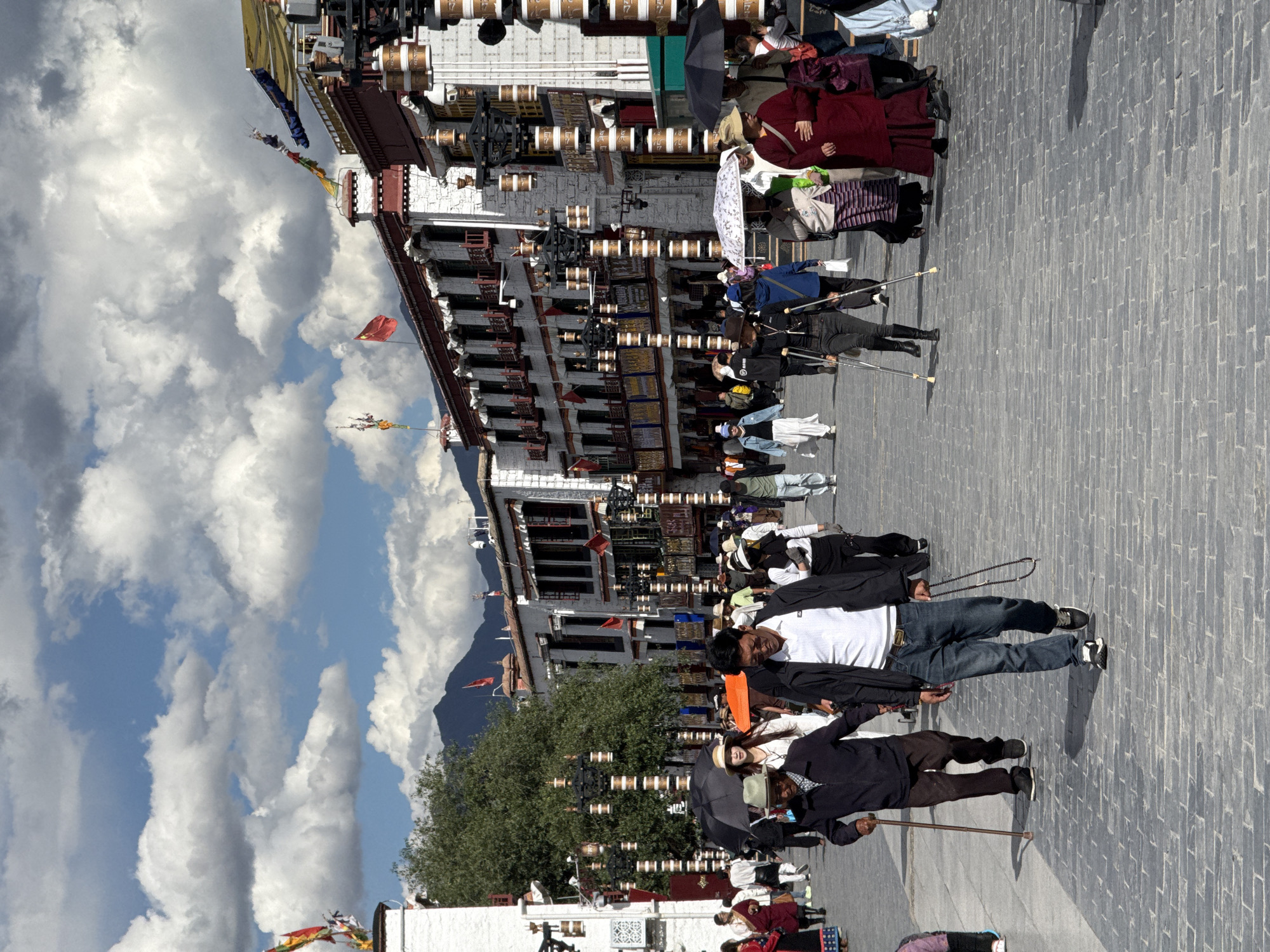 The image captures an outdoor scene, likely a public square or street, in Chengguan District, China. Traditional multi-story Tibetan-style buildings line the left side, characterized by white facades, dark-framed windows, and numerous golden and brown prayer wheels along their structures. Colorful prayer flags are strung above the rooftops, and a red flag flies from one building. A large green tree stands in the mid-ground, and distant mountains are visible under a bright, partly cloudy sky filled with large cumulus clouds.
The paved ground, appearing as gray cobblestone or brick, is populated by many people. Individuals are seen walking and standing, dressed in a mix of traditional Tibetan robes, such as maroon, and modern casual wear including jeans, t-shirts, and jackets. In the foreground, a man in a white t-shirt, blue jeans, and a dark jacket holds a string of prayer beads, while another man in a dark suit jacket uses a cane. Several people are equipped with walking sticks or crutches, and some are holding umbrellas for shade, including one with a distinctive white pattern.
The scene suggests a sunny day, likely late morning or afternoon, given the bright light. The overall activity points to a daily gathering or pilgrimage around a culturally significant site, common in Tibetan areas. No visible text is legible in the image.