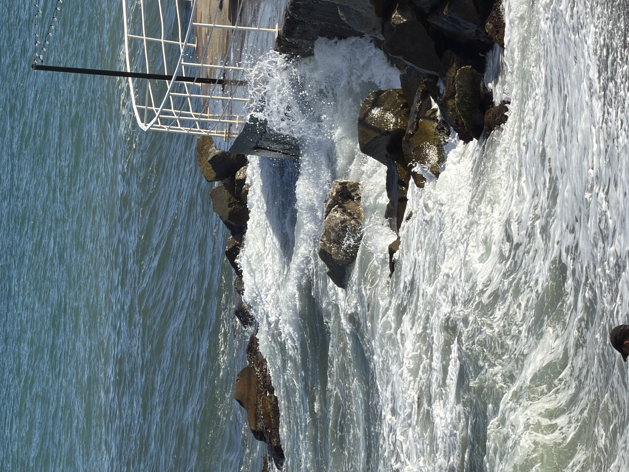A person wearing a dark baseball cap is partially visible in the bottom center of the frame, with their back to the camera. They appear to be standing in shallow water or on a pebbled shore. The image captures a dynamic scene at the Black Sea coast in Alushta, Ukraine. Waves are crashing against a concrete pier and a collection of large, dark rocks. The water is choppy and white with foam, indicating active surf. The pier has a white railing and a tall black pole with string lights extending from it. The sunlight illuminates the water and the wet surfaces of the rocks and pier, creating a bright and textured appearance. The sea stretches out to the horizon, with a clear blue sky above.