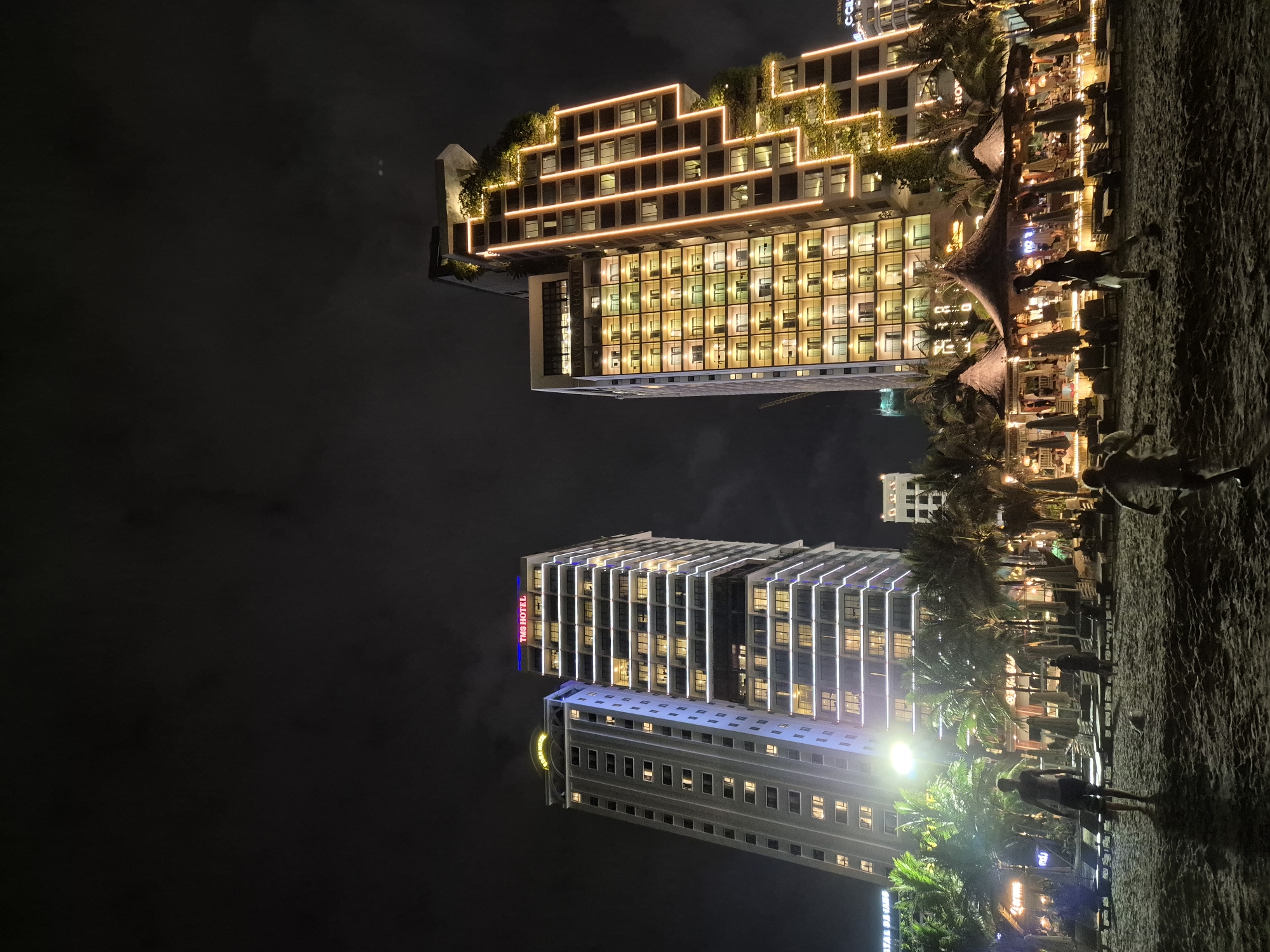 The image depicts a nighttime scene on a sandy beach in Đà Nẵng, Vietnam, with two prominent, illuminated hotels dominating the background. The hotel on the left is a tall, grey building with numerous lit windows, bearing a blue and red sign that reads "TMS HOTEL" at its summit. To its right stands another skyscraper, its facade adorned with vertical orange lights that accentuate its stepped architectural design. This building also features extensive greenery integrated into its structure, particularly on its upper levels.
In the foreground, the beach is populated by several figures. Two young boys are visible playing on the sand, with one actively running. Adults are also present, with one person standing and looking towards the hotels, and another walking in the middle ground. Beach chairs and umbrellas are arranged in front of thatched-roof structures that appear to be part of a beachside establishment, likely a restaurant or bar, also illuminated. Palm trees are scattered throughout the scene, adding to the tropical atmosphere. The night sky is dark and cloudy.