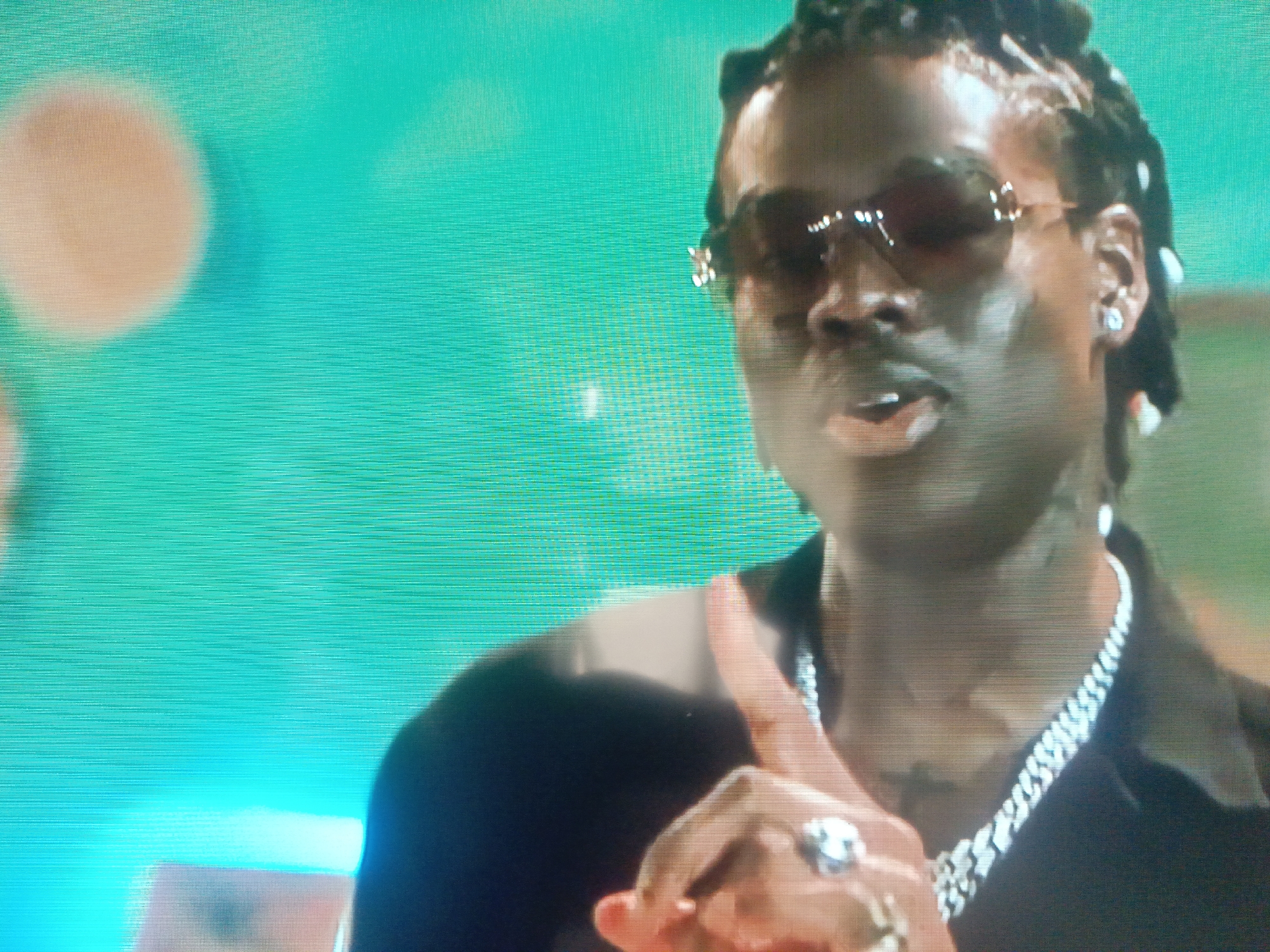 A man with dreadlocks wearing sunglasses, a silver chain necklace, and a ring is in the foreground. His right hand is raised with his index finger pointing up. The background is a solid green color with a blurry, pale circle in the upper left.