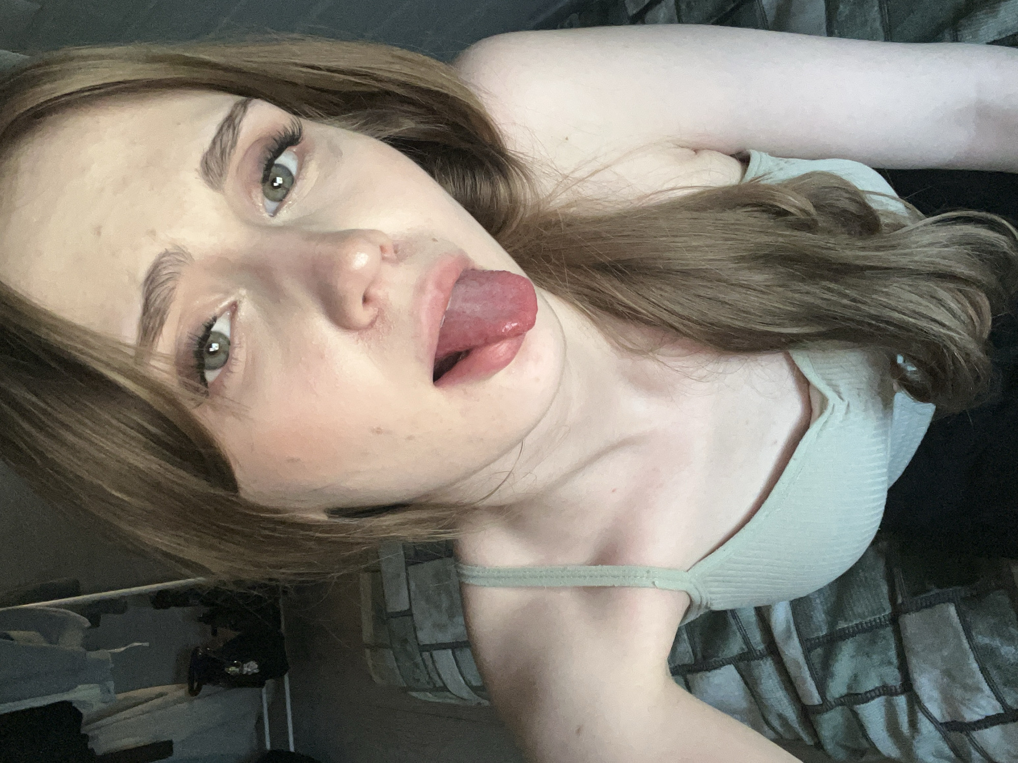 The media file shows a close-up, high-angle, horizontal shot of a young woman with long, light brown hair. She is lying down, with her head tilted back and her tongue sticking out, her eyes looking slightly upward. She is wearing a light green ribbed tank top. The background is dark and out of focus, but appears to be a bed or couch with a patterned blanket. There are some blurred objects in the lower left corner that might be clothing or accessories. The lighting is soft and natural, suggesting it might be daytime. The overall mood is playful or flirtatious. There is no visible text in the image. The location context is Ashkadar, Russia.