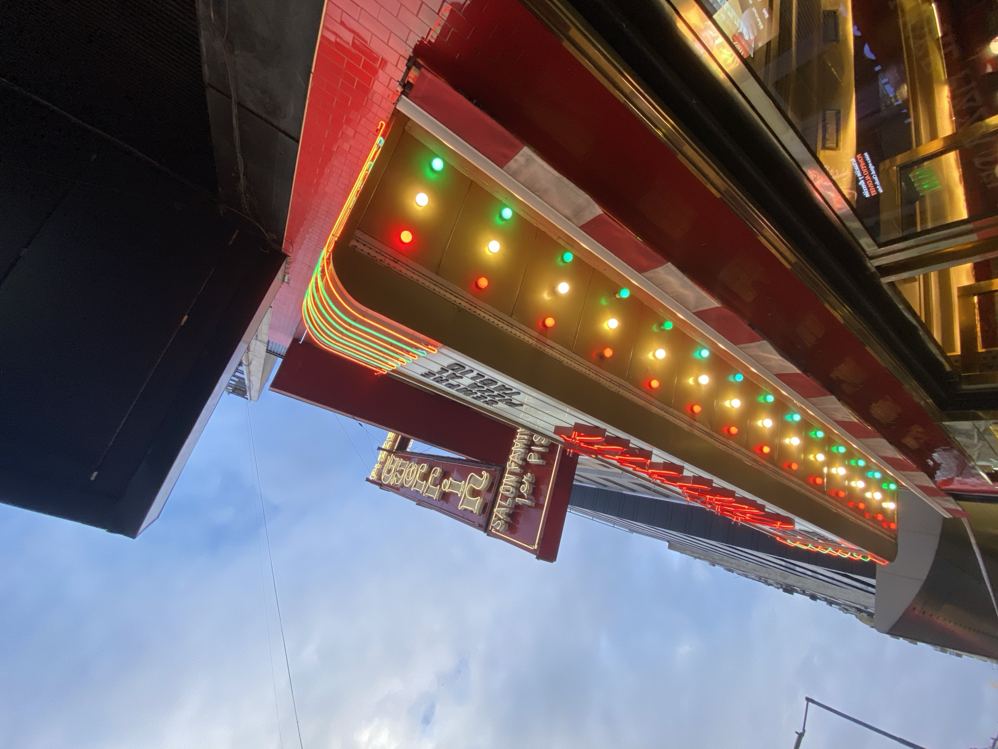 The image depicts a low-angle view of the illuminated facade of Pizzeria Guerrin, located at Av. Corrientes 1368, Buenos Aires, Argentina. The prominent feature is a multi-part marquee and signage system. A vertical sign, cantilevered from the building, displays "PIZZA" and "Guerrin" in yellow neon letters, with "SALON FAMILIAR 1er PISO" in white neon below, all set against a dark red background framed by a red border. Below this, a horizontal marquee features a grid of alternating red, yellow, and green circular lights illuminating a white surface. Above the marquee, the building facade is clad in red tiles. A red and white striped awning extends beneath the horizontal marquee, partially obscuring the upper section of a large glass window. The windows reflect interior lights and indistinct shapes. To the left, a dark, multi-story building rises against a cloudy, light blue sky. Overhead wires are visible. A partial view of a black street light pole is present on the far left. No individuals are explicitly visible.