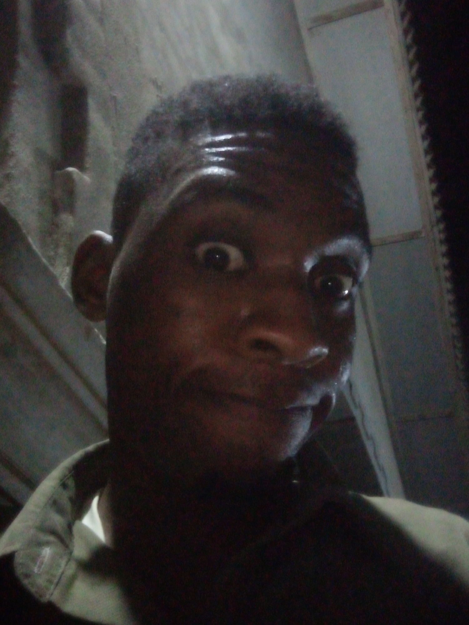 A selfie of a Black man with short, dark hair is shown from a low angle, in a poorly lit indoor location. The man is wearing a dark shirt with the collar visible. His expression is one of surprise or mild amusement, with his eyes widened and a slight smile. The background consists of what appears to be a wall with an indistinct texture. It's not possible to ascertain the precise location or city from this image.