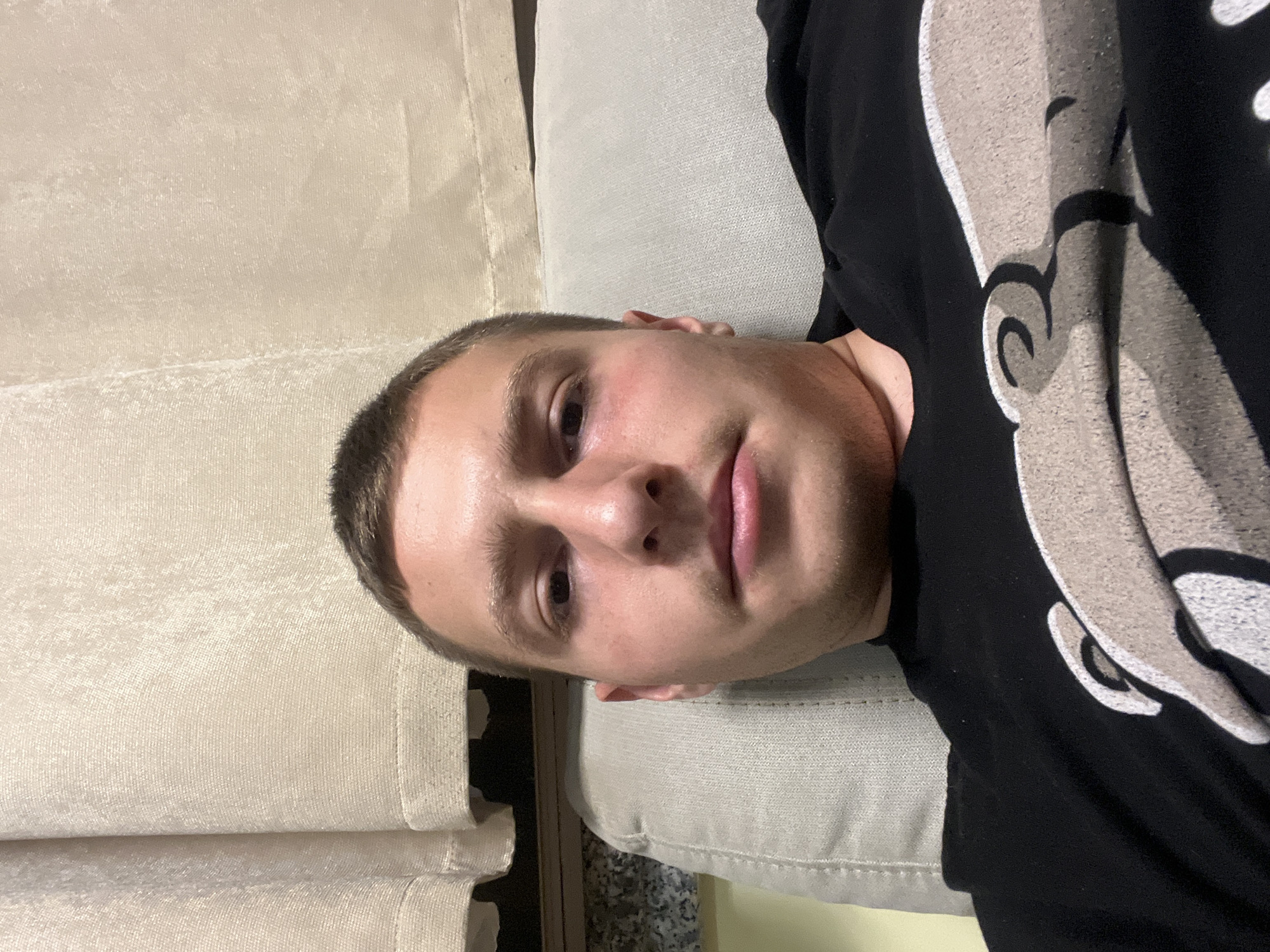 A light-skinned male with short brown hair is lying down, facing the camera. He is wearing a black t-shirt with a light-colored design on it. He is lying on a light-colored, textured surface, possibly a couch or bed. To the left is a light beige curtain. The environment appears to be indoors, likely a residential setting. There is no indication of this image being in Burgas, Bulgaria.