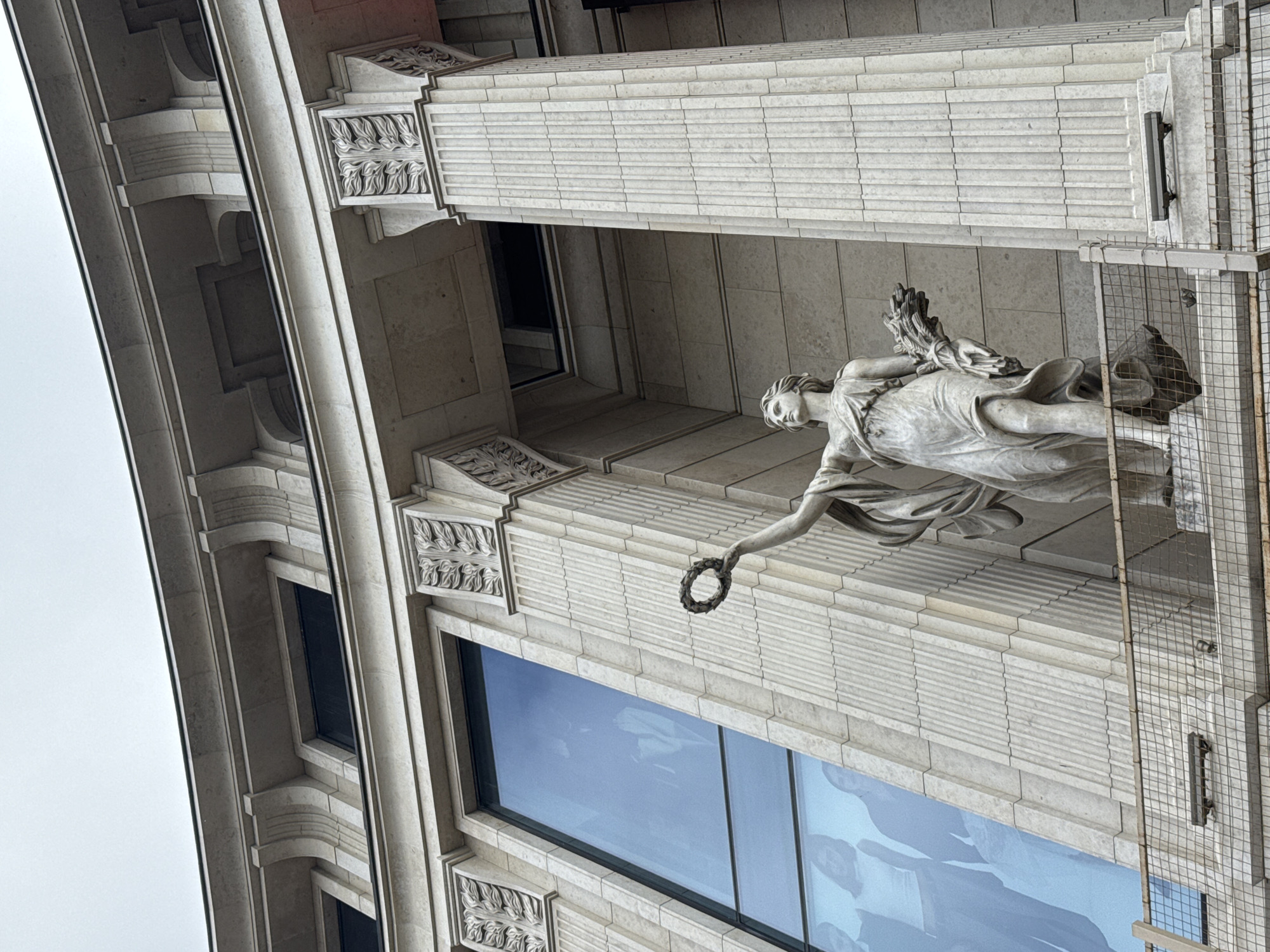 An upward, low-angle, full shot captures a classical-style sculpture of a woman standing on a pedestal outside a building. The sculpture is made of light-colored stone and depicts a woman with her hair pulled back, wearing a draped gown. She is holding a wreath in her raised right hand and a bundle of wheat in her left.
The building behind the sculpture is constructed of light-colored stone with a neoclassical architectural style. Large, rectangular windows with a blue tint are visible, reflecting the overcast sky. The building features fluted columns, ornate carvings above the windows, and a curved roofline. A metal fence or railing runs across the bottom of the image, in front of the pedestal. The overall scene suggests an outdoor public space in a city.