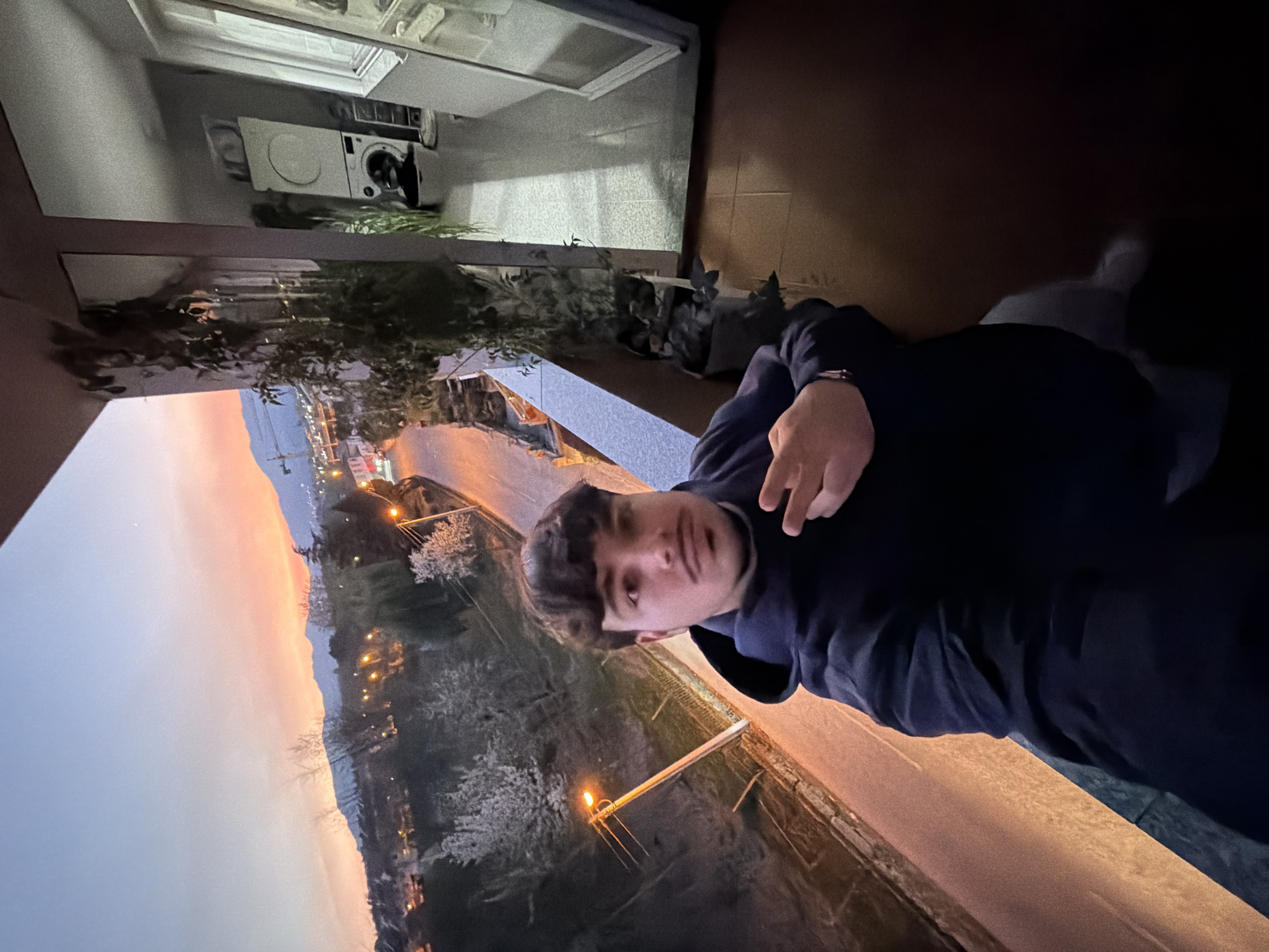 A person is on a balcony with a view of Vila Real, Portugal at sunset. The sky shows a gradient from blue to orange. In the foreground, there is a railing, and blooming trees can be seen. The person is wearing a dark hooded sweatshirt and has dark hair. A washing machine is visible through a window. The person is making a hand gesture. The balcony has tile flooring. Streetlights illuminate the cityscape.