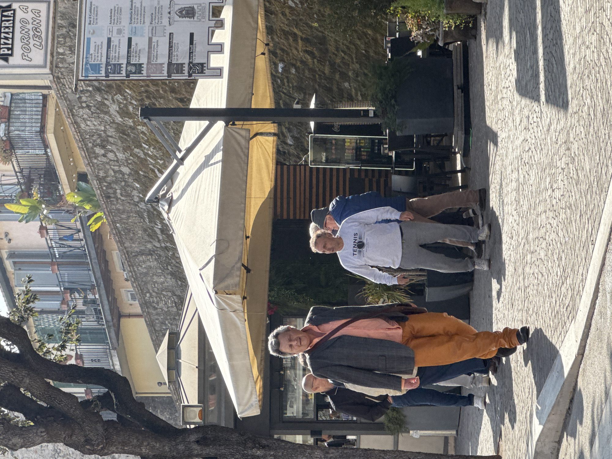 The image shows a street scene in Nettuno, Italy. A group of people, mostly men, are standing on a cobblestone street next to outdoor seating. One man wears orange pants and a grey suit jacket over a salmon-colored shirt. Another man is wearing a sweatshirt with the word "TENNIS" printed on it. The scene is located next to a building with a sign that reads "PIZZERIA FORNO A LEGNA." Several nearby establishments are named: "Bar il Gabbiano," "Accademia," "Trattoria Romolo," and "Pizzeria L'Archetto." Coca-Cola-branded refrigeration units are visible in the background. The setting appears to be a commercial area with restaurants and shops. A tree occupies the lower left foreground.
