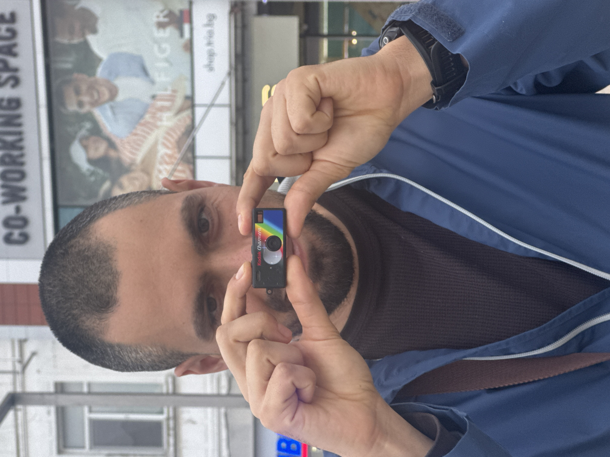 A man with short, graying hair and a beard holds a black Kodak Charmera mini camera in front of his face, framing it between his thumb and index finger. He is wearing a dark gray shirt under a blue jacket. On his left wrist is a black digital watch. The background features a street scene with a blurred storefront sign displaying the words "CO-WORKING SPACE" and part of a Tommy Hilfiger advertisement. The location is indicated by the website "shop.tria.bg" visible on the advertisement, placing the scene in Burgas, Bulgaria. A partial neon sign is visible at the bottom of the frame.