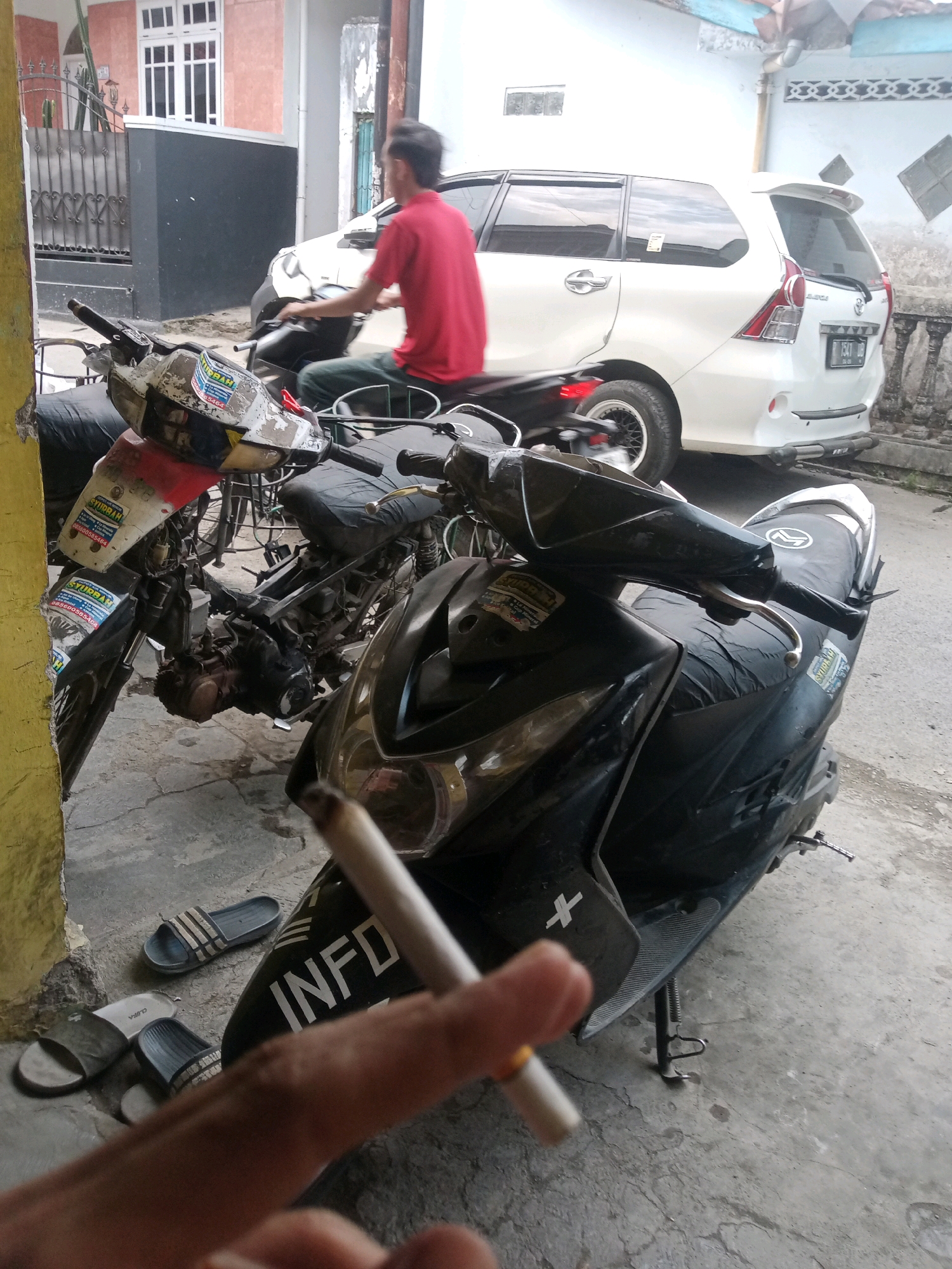 The image depicts a street scene in Grendeng, Indonesia, focusing on several motorcycles parked on a paved area. In the foreground, a person's hand holds a lit cigarette, partially obscuring the view of a black scooter. The scooter has "INFO" written in white on its side. Next to the scooter are two pairs of flip-flops. Behind the scooters, a white van is visible, and a person in a red shirt is riding another motorcycle. The background includes a building with windows and a fence. The overall environment suggests an outdoor, possibly residential or commercial, setting in a town or city.
