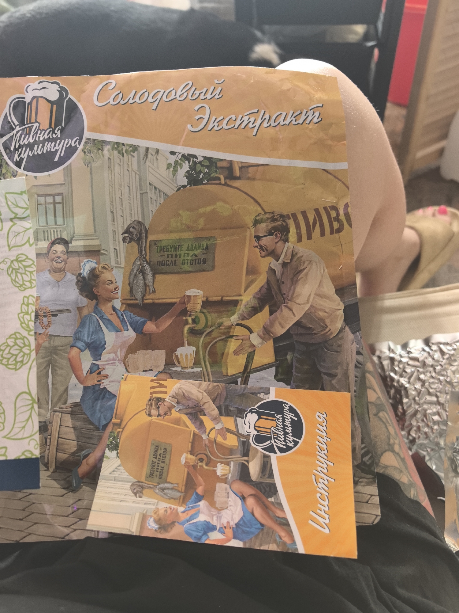 The image displays a printed document, likely product packaging or a brochure for a "Malt Extract" product, held open on a person's lap in an indoor environment. The document features retro-style illustrations and entirely Cyrillic text, including "Солодовый Экстракт" (Malt Extract), "Пивная Культура" (Beer Culture) with a beer mug logo, and "Инструкция" (Instruction). The specific photographic location within the city of Noviy, Russia, is not visually presented in the image; however, the Russian text on the packaging indicates its origin or distribution in Russia.
The central artwork on the document depicts a bustling street scene around a large, yellow mobile beer dispenser. In the upper left, a smiling man holds a string of sausages. To his right, a smiling woman in a blue dress and white apron dispenses beer into a mug from the yellow tank. Above the dispenser, a cat attempts to snatch fish hanging from a hook. Text on the dispenser reads "Требуйте допива пива после отстоя" (Demand a topping up of beer after settling). In the lower, tilted section of the artwork, another woman in a blue dress and white apron is shown reclining on a lower part of the dispenser, holding a beer mug, while a man in glasses is bent over the machinery, seemingly operating it.
The immediate environment of the photograph shows a light-skinned arm and knee of the person holding the document, with dark fabric (likely trousers or shorts) underneath. A light-colored slipper is visible on one foot, revealing toenails painted pink. To the right of the person's leg, a crumpled piece of aluminum foil is present. The background is an out-of-focus indoor setting, featuring dark furniture, a red object, and light-colored wooden paneling.