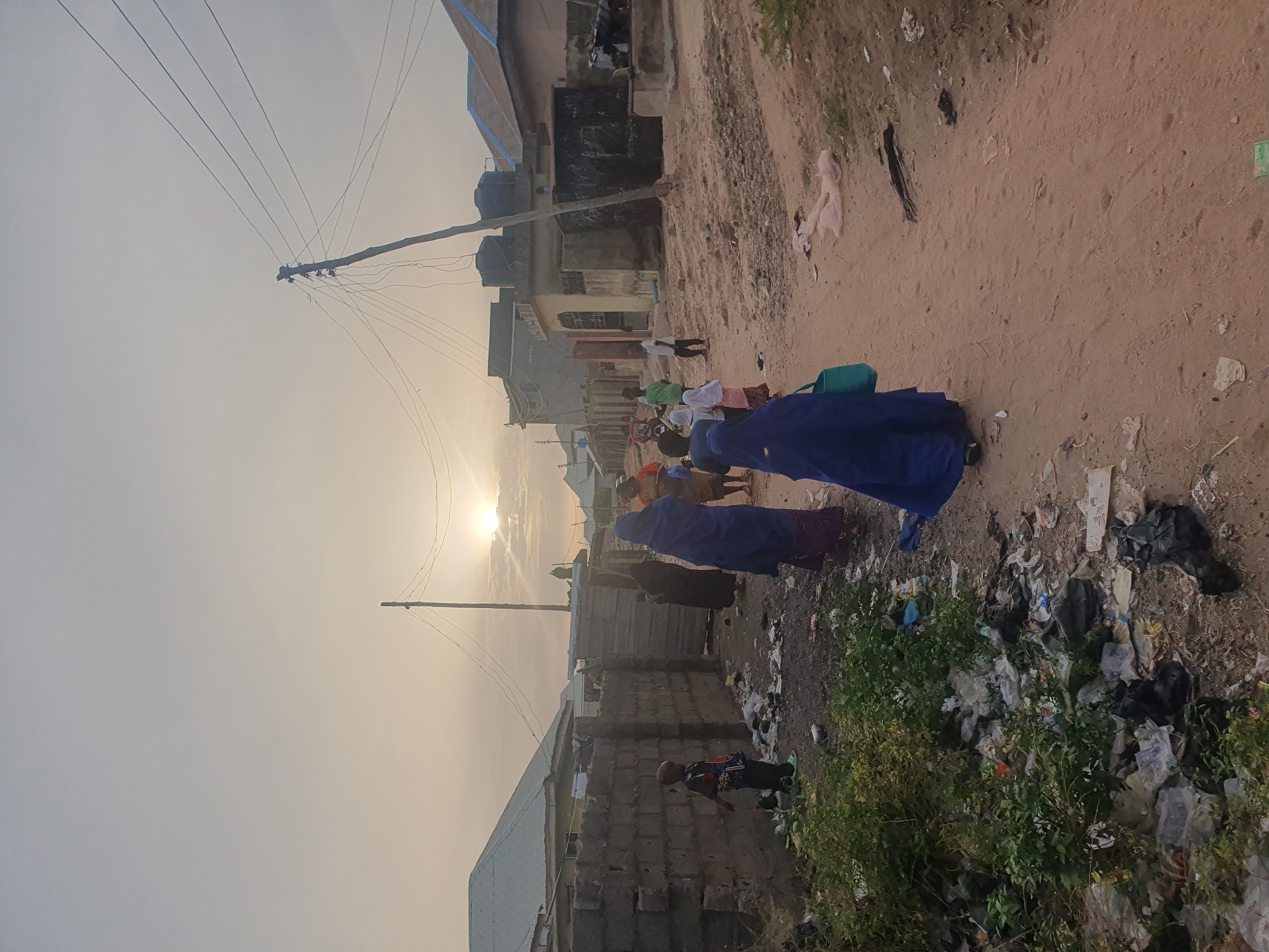 The image depicts an unpaved street in Lokoja, Nigeria, at dusk or dawn. Power lines stretch across the sky. People in various clothing are walking on the street. A woman wearing a blue garment appears prominently in the foreground. Piles of trash litter the edge of the road, near vegetation. Buildings, some seemingly made of cinder blocks, line the other side of the street. A child is on the right side of the photo, near the garbage and vegetation.