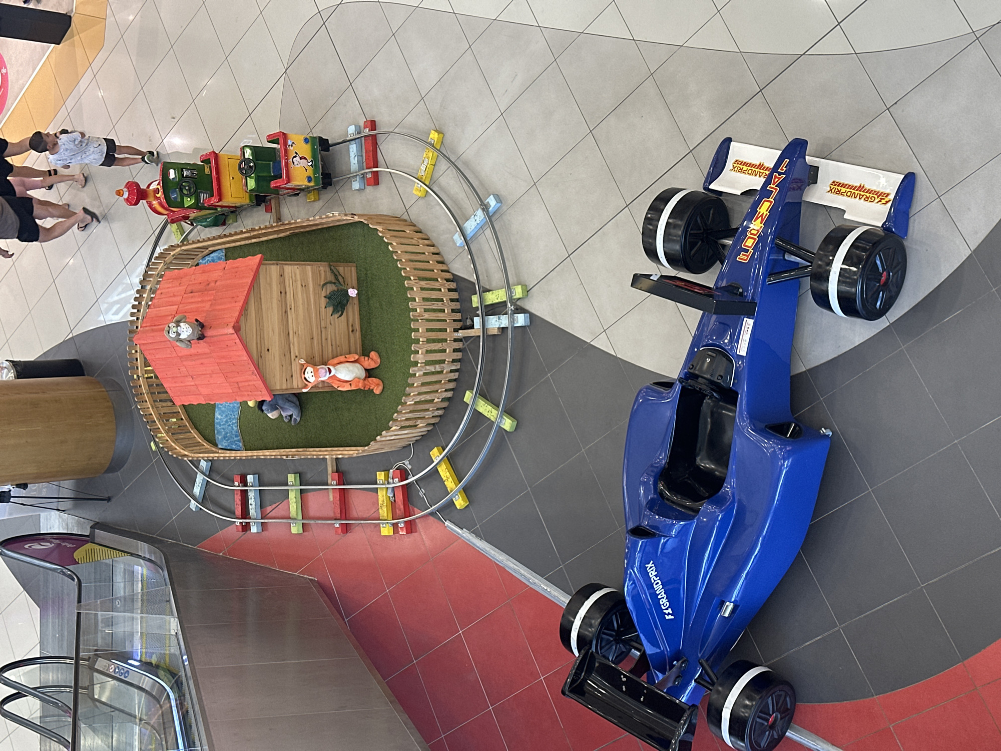 The image depicts a scene from an indoor location, possibly a shopping mall, as suggested by the presence of an escalator in the background and tiled flooring. In the foreground, a blue ride-on race car with "F1 GRANDPRIX" branding is positioned on the floor. Behind the car, a miniature train track circles a small, wooden playhouse with a red roof. The train consists of a red and green engine and several colorful carriages, some with cartoon figures. A stuffed Tigger toy and a stuffed owl are placed near the playhouse. Two children are visible in the middle ground, one walking away from the camera and another walking towards the train. The floor is a mix of red and gray tiles.