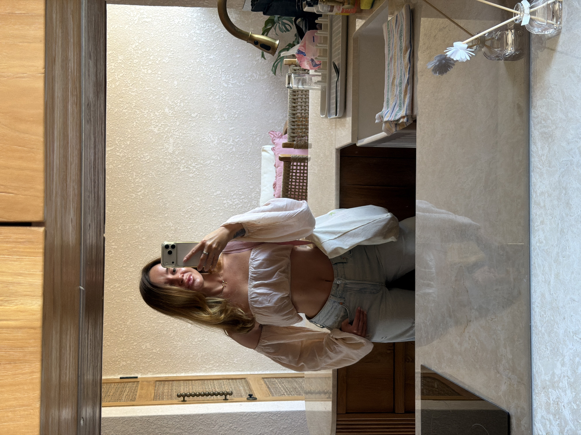 A woman is taking a selfie in a bathroom. She is wearing a light pink off-the-shoulder crop top and light blue jeans. She has on a thin necklace and a ring on her left index finger. She is holding her phone in her right hand. A light pink bag is slung over her left shoulder. In the background, there is a sink, a faucet, a dish rack with glasses and other items, a cabinet, and a doorway. The scene is well-lit, suggesting it is daytime. The woman's expression is playful, with her lips slightly parted.