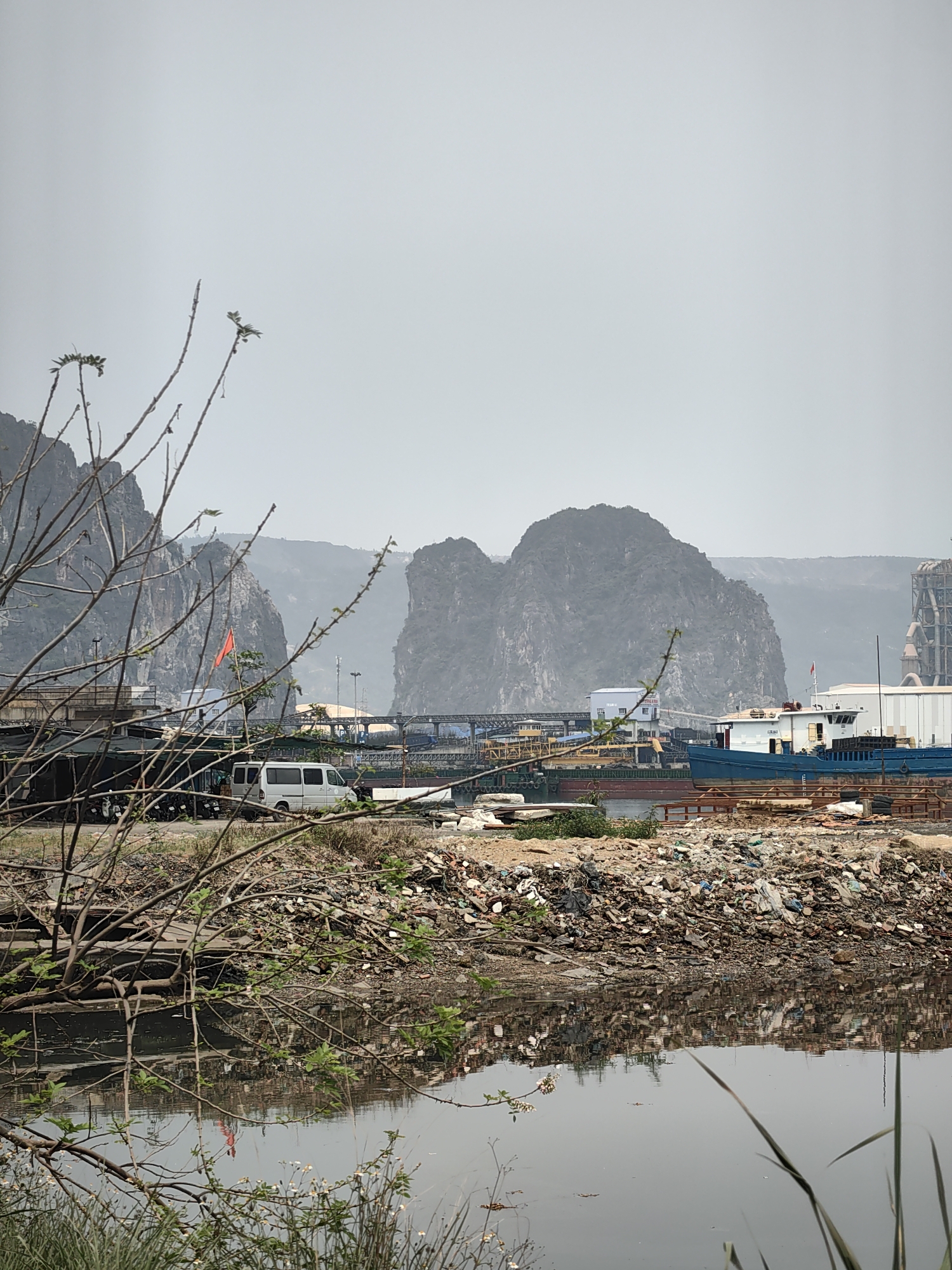 The image depicts a polluted waterway in Quang Hanh Ward, Vietnam. A large pile of trash and debris lines the bank of the water, with a reflection of the debris visible in the water. In the background, there are limestone karsts characteristic of the region, alongside industrial infrastructure including ships and a cement factory. A white van and parked motorcycles are visible behind the trash. A bare tree branch with sparse foliage is in the foreground. The sky is overcast. The overall scene conveys environmental degradation in an industrial area.