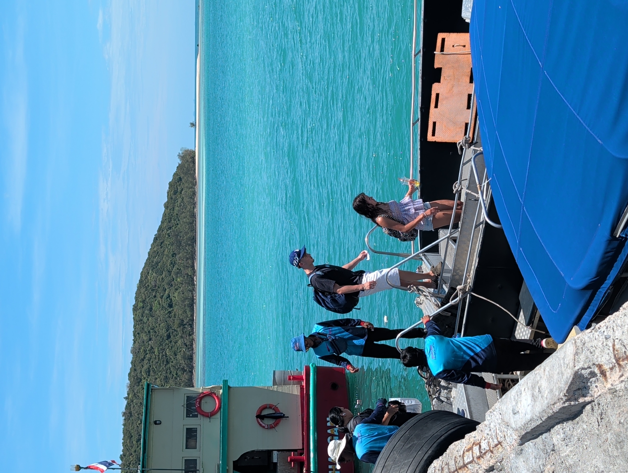 Here's a description of the image:
The image shows people disembarking from a boat onto a pier. The location appears to be in ตำบลเกาะพะงัน, Thailand, given the tropical scenery and geographical location.
There are a few people on the boat, descending a set of metal stairs. One person is wearing a black shirt, white shorts, and a blue baseball cap, carrying a dark-colored backpack. Another person is wearing a light-colored dress and is holding a bottle. Below them are some crew members, wearing blue uniforms.
The boat is docked next to a concrete pier. The background shows turquoise water, a green forested island, and a clear blue sky. There is also a sandy beach visible along the coast of the island. The scene appears sunny and bright.