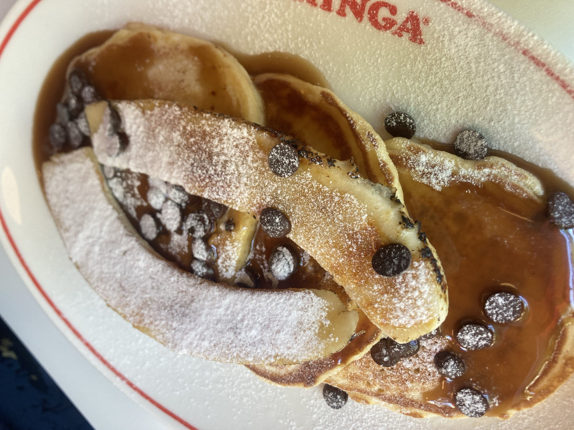 A close-up, top-down shot displays a plate of pancakes, syrup, chocolate chips, and powdered sugar. The pancakes are golden brown and stacked, with two banana slices on top, dusted with powdered sugar. Chocolate chips are scattered across the pancakes and in the syrup. The plate has a red rim and white interior, with the partially visible red text "NGA" from the word "MANGA" printed on it, suggesting the location is a restaurant or cafe named "MANGA" in Barcelona, Spain. The background is dark blue and out of focus.