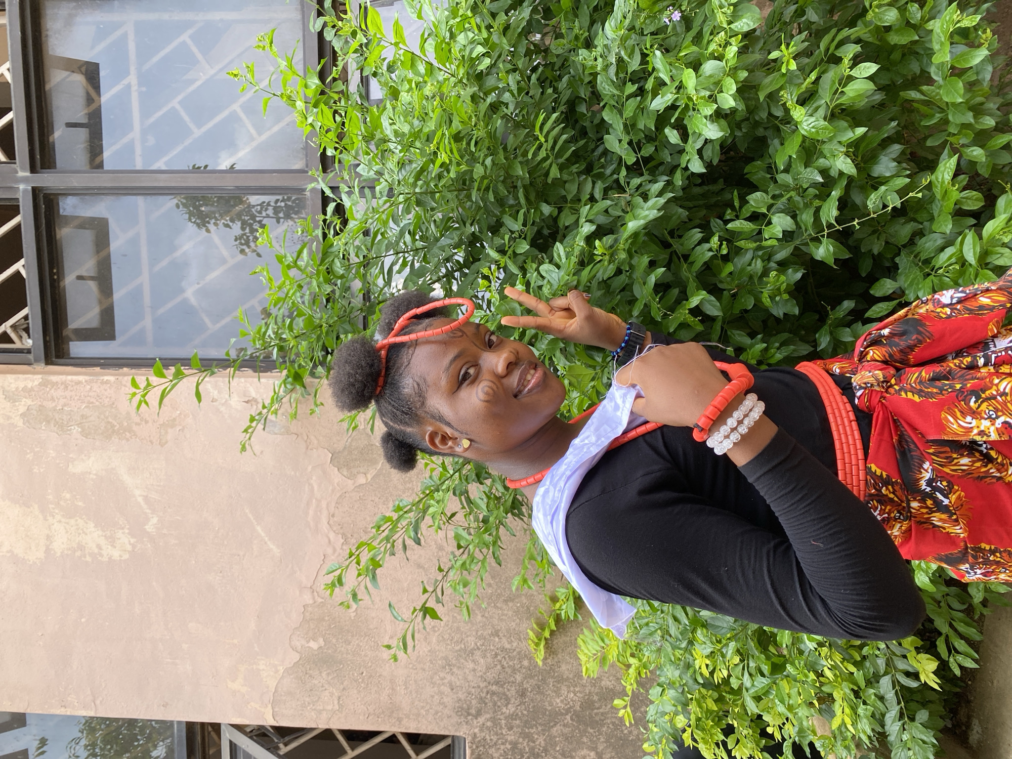 A young woman is posing for a photo outside a building in Gwong, Nigeria. The woman is wearing a black shirt, a red and gold patterned skirt, orange beaded jewelry on her head and neck, and white and orange bracelets on her wrist. She has traditional face paint markings. She is making a peace sign with her fingers. A white cloth is draped over her shoulder. Behind her is a green leafy bush and a building with windows. The wall of the building is beige with some peeling paint.