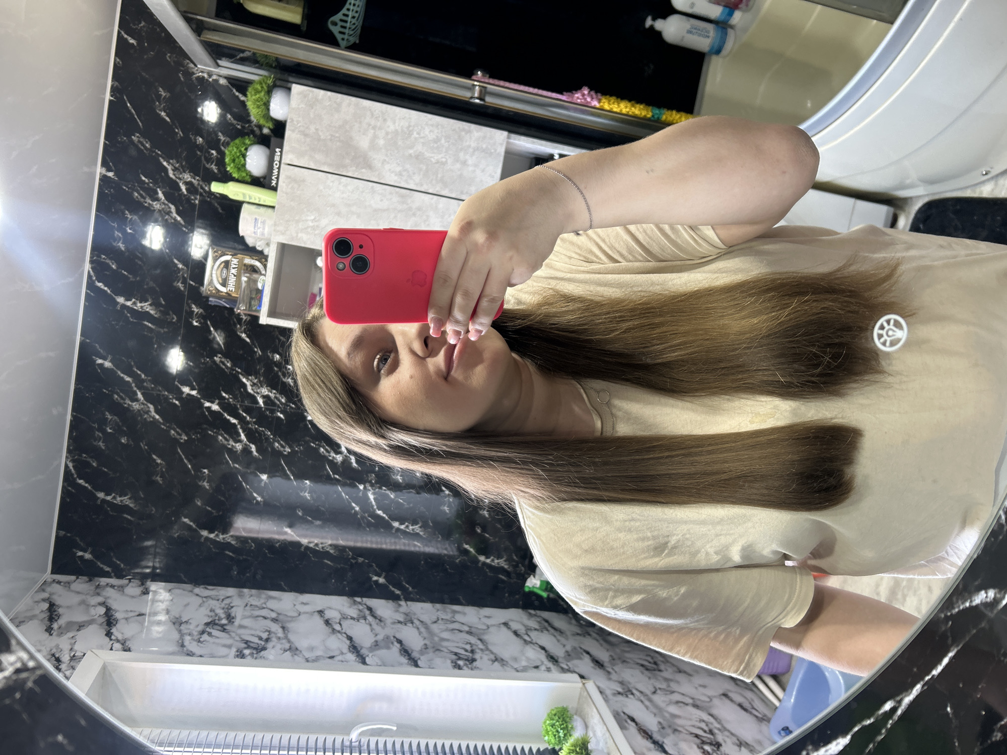 A person with long brown hair is taking a selfie in a bathroom. The bathroom features black and white marble-patterned walls and countertops. Various toiletries and decorative plants are visible on the shelves and counters. The location is Ольховка, Russia.