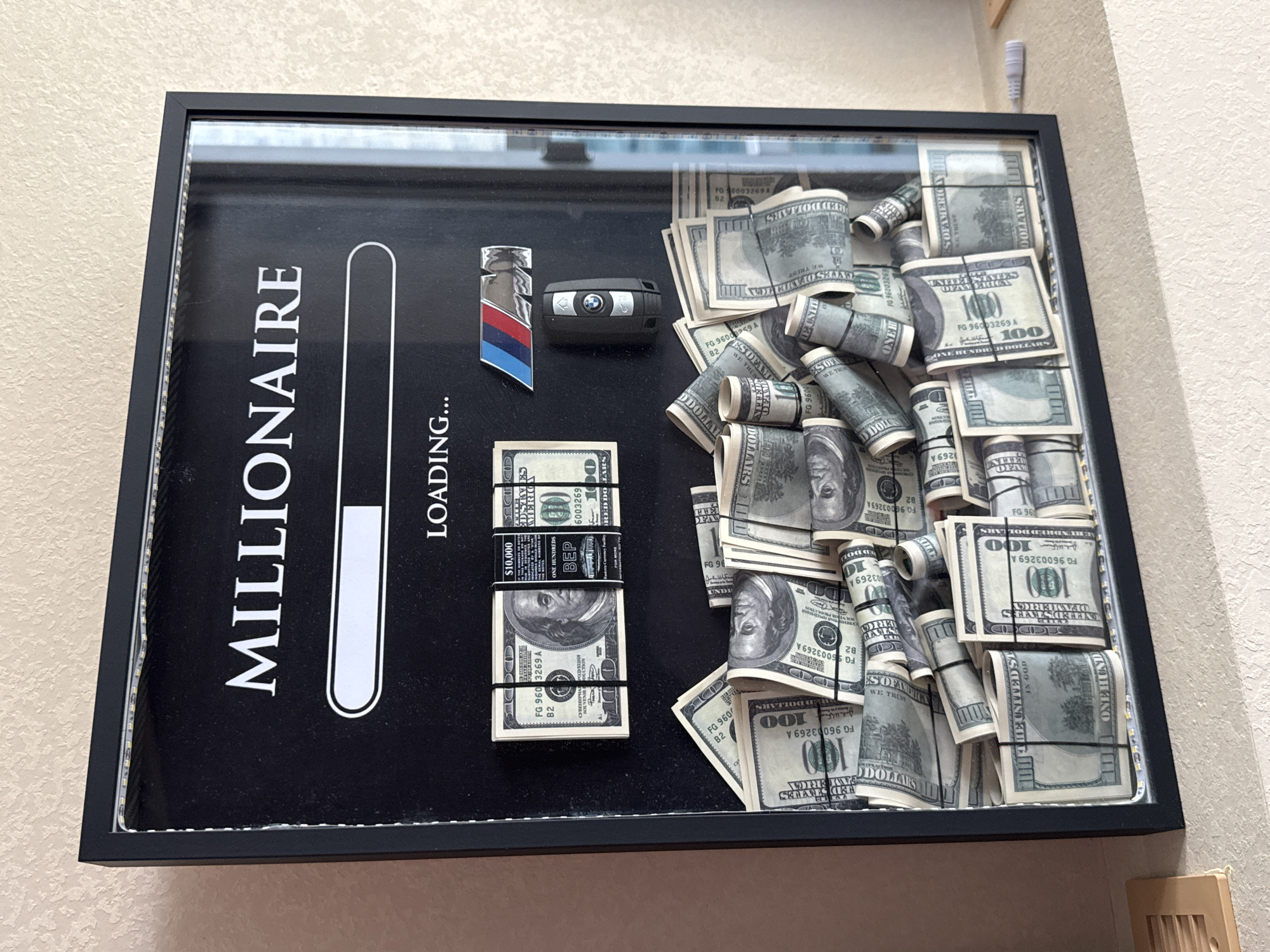 The image shows a shadow box frame displayed indoors, likely on a shelf or mantlepiece, against a textured wall. The shadow box is black with a glass front. Inside, the top of the frame features the word "MILLIONAIRE" in white capital letters, followed by a progress bar that is partially filled. Below the progress bar are the words "LOADING..." and an emblem that appears to be a BMW logo, along with a BMW car key. The majority of the shadow box is filled with numerous stacks and rolls of US dollar bills, specifically $100 denominations. These bills are bundled with black rubber bands. Some bundles are stacked neatly, while others are loosely arranged, creating a visually dense display of money. The bottom edge of the shadow box is illuminated by white LED strip lights. There are no people or animals visible in the image, and no specific geographical markers indicating a location in Yekaterinburg, Russia. The environment appears to be a domestic or office setting.