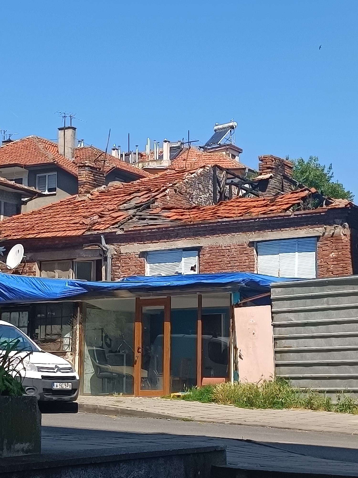 The image captures a damaged building with a partially collapsed terracotta-tiled roof under a clear blue sky. The brickwork is exposed and in disrepair in several areas. A blue tarp is stretched over a lower section of the building, which appears to be a commercial space with large glass windows. Inside the commercial space, furniture, including chairs, is visible, suggesting it might be a salon or shop. A white van with a license plate "A 5079 PA" is parked in front of the building. To the right of the commercial space, a corrugated metal fence is erected, and there is an overgrown patch of grass. The scene appears to be on a street with a paved sidewalk. In the background, other buildings with similar tiled roofs and chimneys are visible, along with a solar water heater on one of the roofs. The overall impression is one of a street in an urban area, possibly showing the aftermath of an incident that caused structural damage to the building. The location is identified as Burgas, Bulgaria.