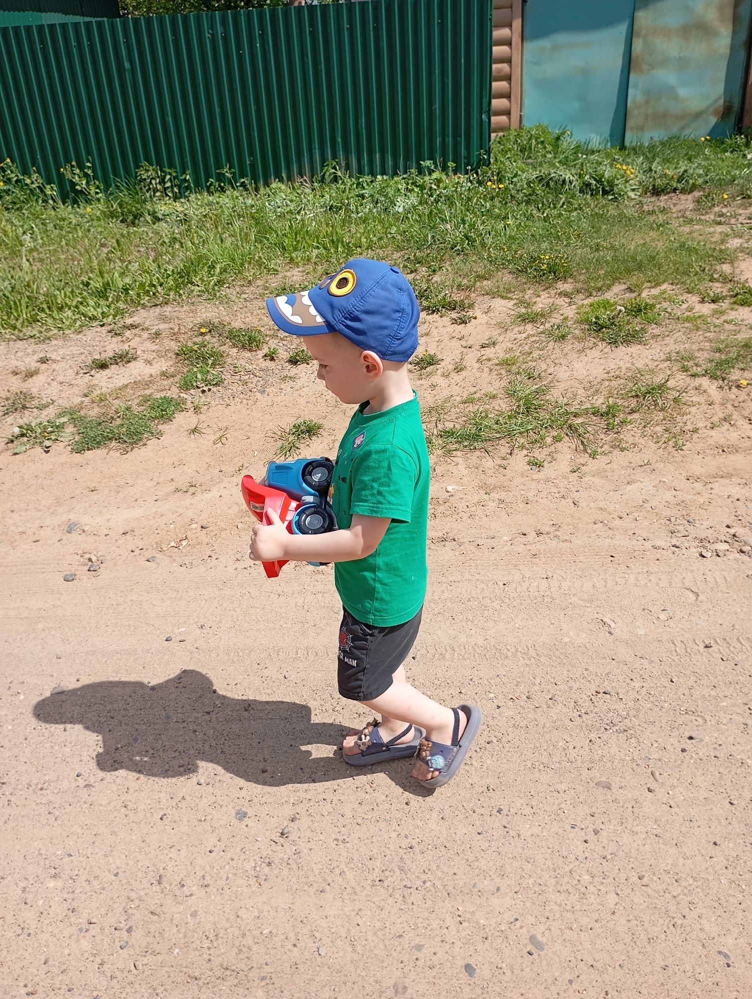 A young child is walking on a dirt path in an outdoor setting. The child is wearing a green t-shirt, black shorts with "Spider-Man" on the left leg, and sandals. He has a blue cap with a yellow circular design. He is holding a red and blue toy truck. The path appears to be gravel and dirt, with sparse green vegetation on either side. In the background, there is a green corrugated fence, with a section of wooden structure attached on the right. The shadow of the child is visible on the path. The location and city are not discernible from the image.