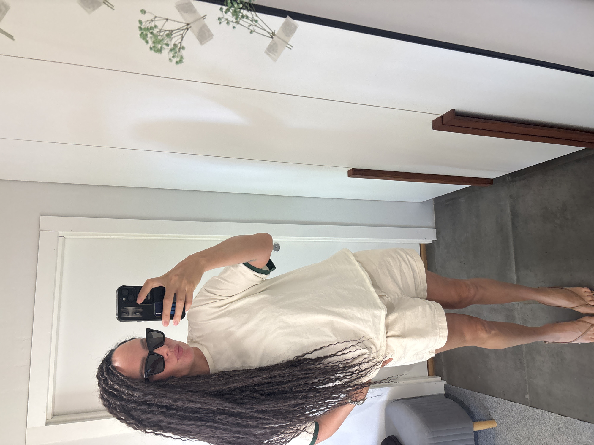 The image depicts an indoor scene in the city of Chaikovsky, Russia. A female individual is positioned in front of a mirror, capturing a self-portrait with a smartphone. The individual has long, dark, braided hair that extends past her waist. Her face is partially obscured by dark, rectangular-framed sunglasses and the smartphone. She is attired in a loose-fitting, light beige short-sleeved t-shirt with dark green piping on the sleeves and matching light beige shorts. She wears thin-strapped, light brown flip-flops on her feet, with dark-colored polish visible on her toenails. A silver-toned watch is present on her left wrist. The individual holds a black smartphone, encased in a rugged black cover with an attached card holder displaying a blue card, in her right hand, while her left hand rests on her hip.
The reflective surface is a large, full-length mirror. In the reflection, a white door frame is visible on the upper left, and a grey upholstered cylindrical stool with light wooden legs is partially visible on the lower left. The floor, both in the reflection and the unreflected foreground, consists of large, dark grey tiles or concrete. To the right of the mirror, a white wall is adorned with several small clusters of green foliage, each secured with light-colored adhesive tape. Two vertical dark brown wooden elements, appearing as frames or trim, are leaning against the wall in the lower right section of the image. The lighting is uniform, indicating an interior setting.