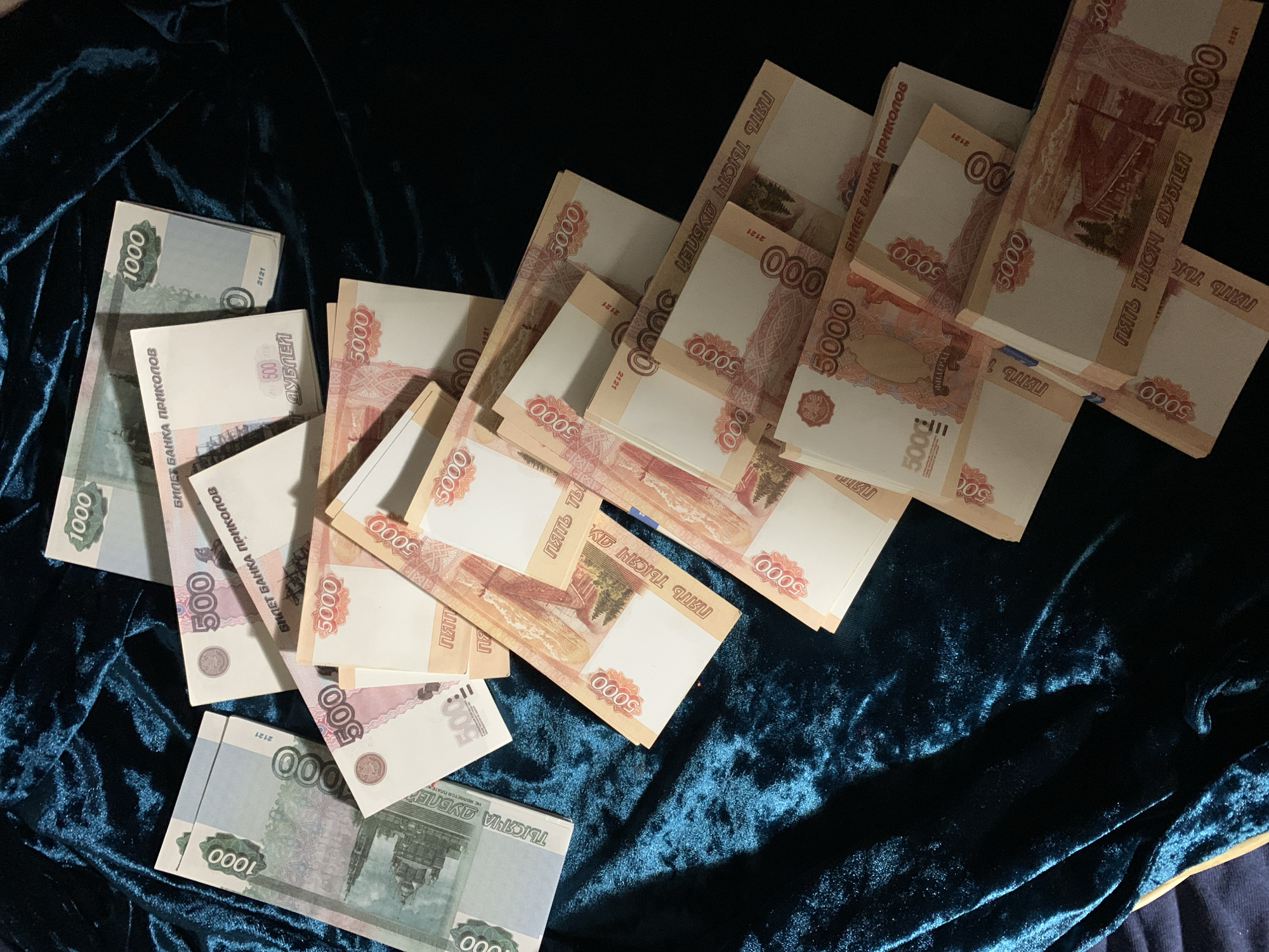 The image shows a collection of play money scattered on a dark teal velvet surface. There are stacks of 5000 "дублей" (rubles), and loose bills of 1000 and 500 "дублей" visible. The bills are marked "БИЛЕТ БАНКА ПРИКОЛОВ," indicating they are souvenir or novelty items. There are no people visible in the image, the location is undeterminable.