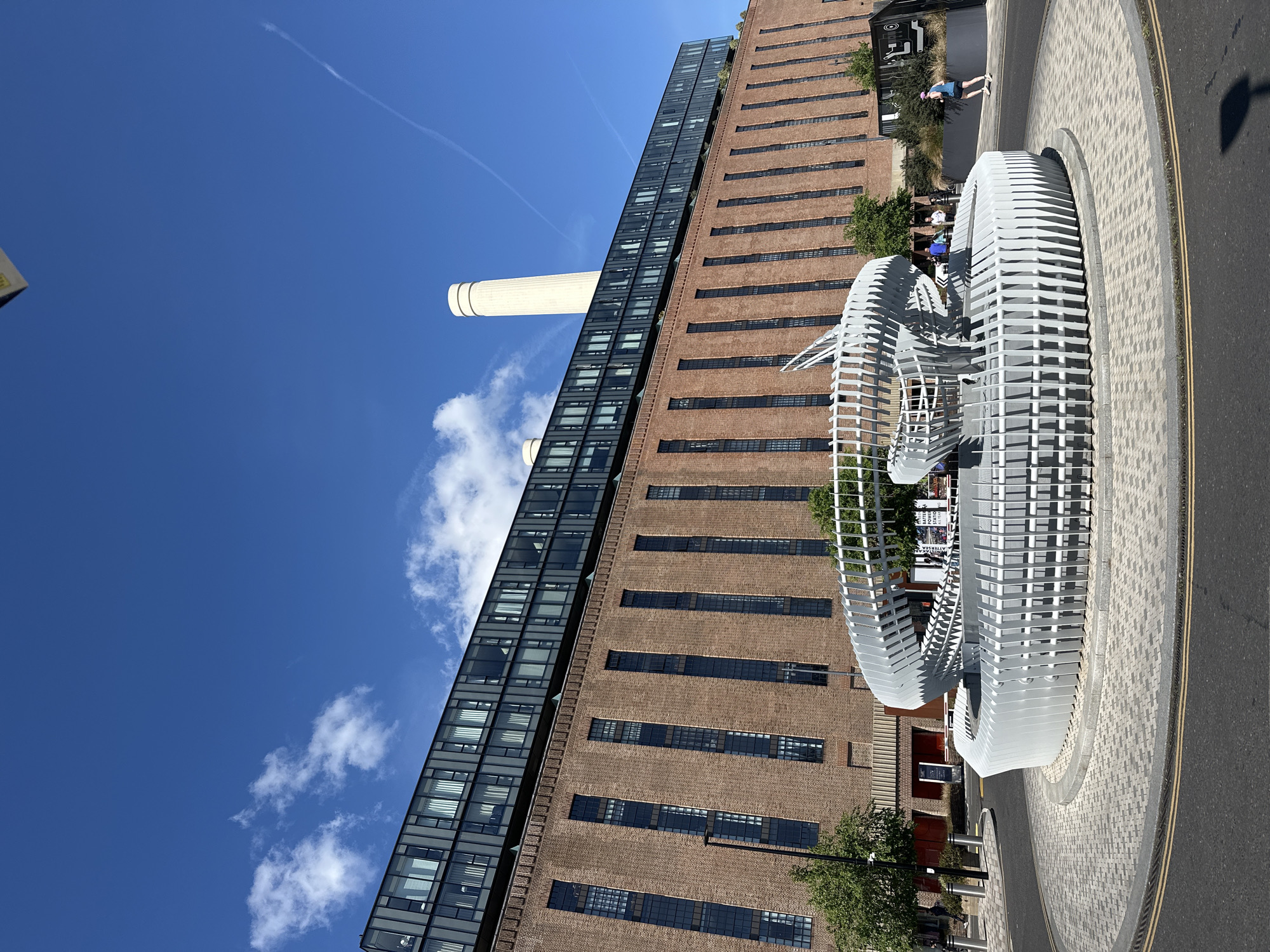 The image features the Battersea Power Station in London, United Kingdom, under a bright blue sky with a few scattered white clouds. The power station, a large brick building with prominent chimneys, is seen alongside a modern glass-fronted structure. In the foreground, there is a large, sculptural installation composed of white, curved metal elements. A paved roadway borders the installation. A few people are visible near the base of the building, some sitting and others walking.