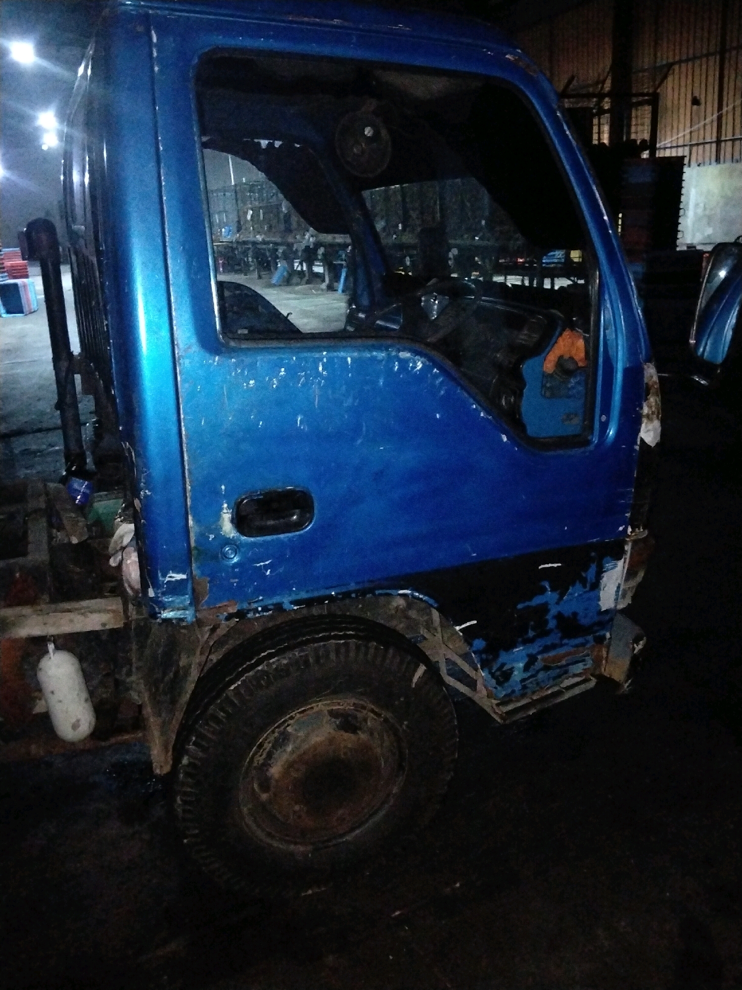 This is a medium shot, eye-level, medium-quality, dimly lit photograph of the side of a blue truck. The truck is in a dark, indoor environment that appears to be a workshop or storage area. The background shows some industrial elements like metal shelving and what looks like other vehicles or large objects. The truck itself is worn, with visible rust and damage to its blue paint, particularly on the lower front section. A black tire is prominently visible on the lower right. Inside the truck's cabin, the steering wheel and dashboard are partially visible. A white cylindrical object, possibly a tank or float, hangs from the side of the truck's chassis near the front wheel. The lighting is artificial and appears to be overhead, casting some light on the truck but leaving much of the environment in shadow. There are no people clearly visible in the immediate vicinity of the truck. The setting suggests a location in the industrial or commercial sector of Moro, Indonesia, likely where vehicles are repaired, stored, or sold.