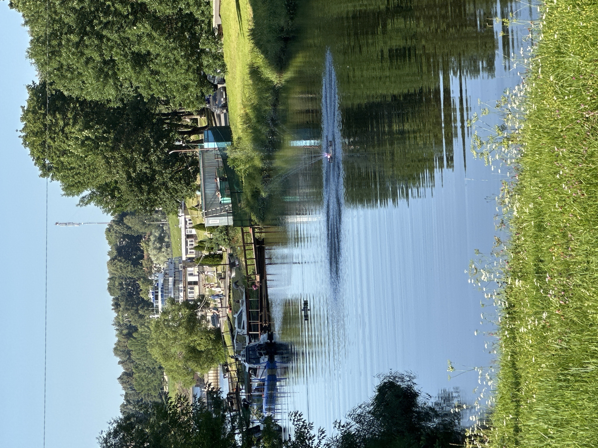 This image shows a wide, calm river with a small, remote-controlled boat creating a spray of water in the center. The riverbanks are lined with lush green grass and wildflowers in the foreground. In the mid-ground, there are several docked boats, including a white passenger boat and a smaller blue and white vessel. A wooden pier extends into the water. Beyond the river, a green grassy slope leads up to a road where a few cars are parked. In the distance, a large white building with multiple levels and balconies is visible, surrounded by trees. The background is dominated by a dense forest and a clear blue sky. A tall telecommunications tower stands on a distant hill. The scene appears to be a recreational area in or near the city of Selivanovo, Russia.