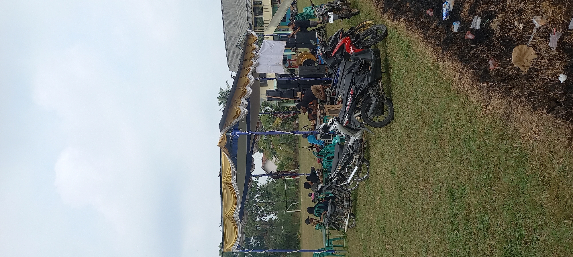 The image depicts an outdoor event set up in a grassy field, likely in Sidamulya, Indonesia. A temporary stage structure, covered by a yellow and white canopy supported by blue and white poles, is prominent. Underneath the canopy, a sound system with multiple speakers is set up, and several people are interacting with equipment, suggesting a musical or performance event. A white banner is also displayed. Several motorcycles are parked in the foreground and mid-ground. In the background, a building with a gray roof and light-colored walls, possibly a school or community hall, is visible. The environment is a grassy field with some trees and a soccer goal in the distance. Scattered on the ground near the foreground, there is some debris, including plastic cups and burnt material, indicating a recent fire or cleanup. The sky is overcast.