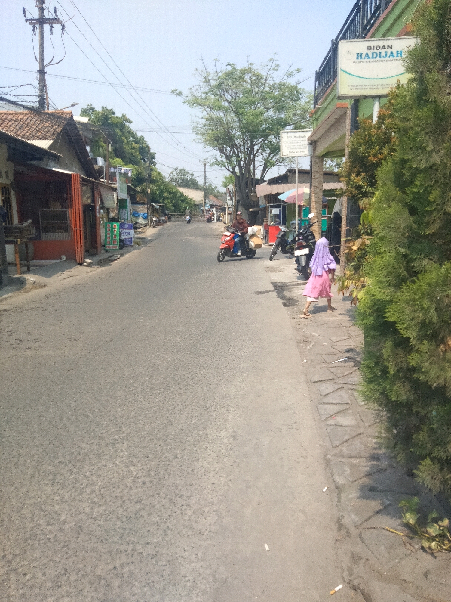 The image depicts a street scene in Pisangan Jaya, Indonesia, on a sunny day. The road is paved, with a sidewalk on the right side. A young child wearing a pink dress and a light purple headscarf is walking on the sidewalk. To the left of the road, there are several small shops with orange and brown storefronts. Across the street, on the right, is a building with a green and white sign that reads "BIDAN HADIJAH". Several motorbikes are parked on the side of the road. In the distance, more buildings and trees line the street. The sky is clear and bright.