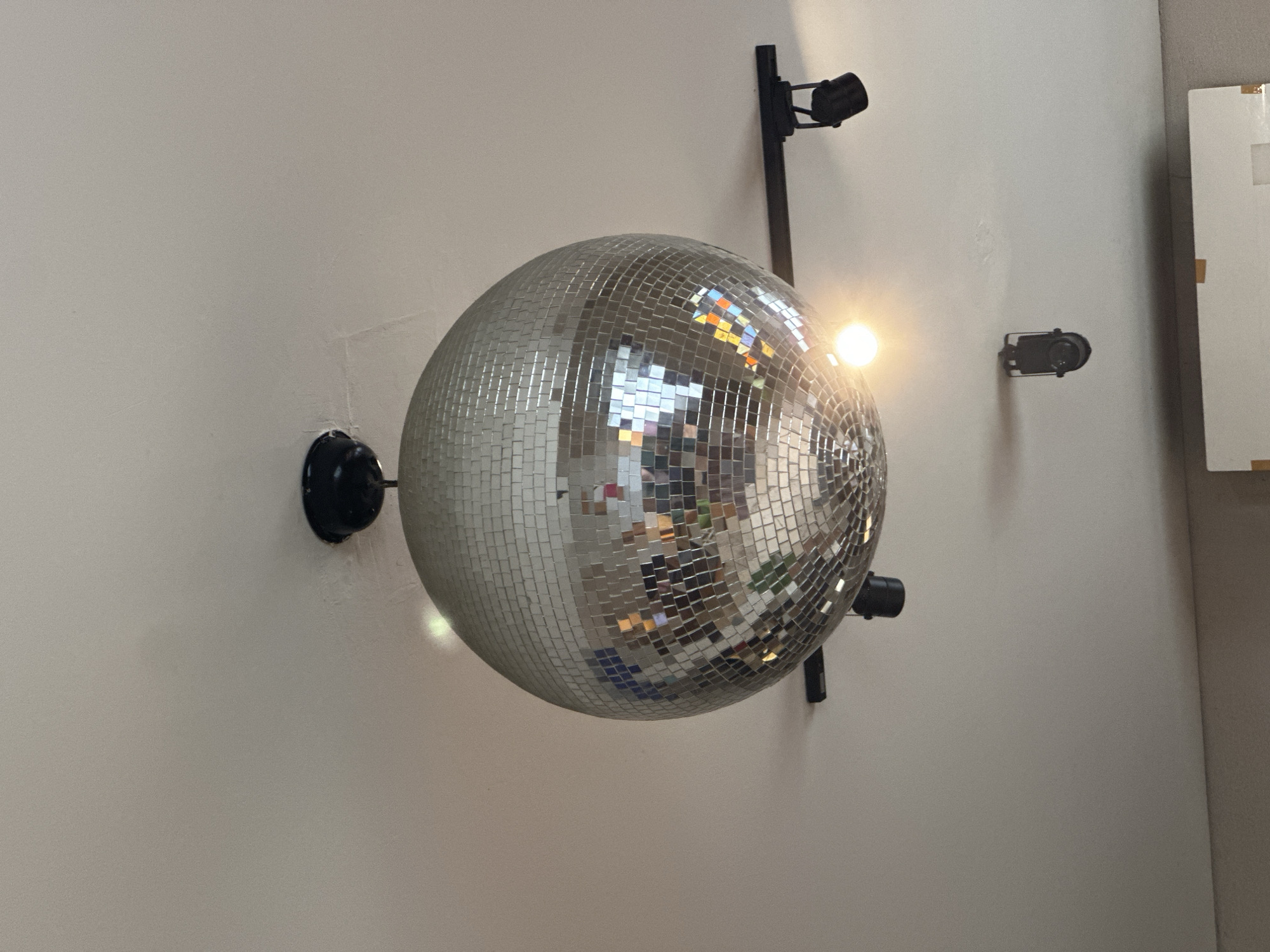 A large, spherical disco ball, covered in numerous small, square mirror tiles, dominates the center of the frame. It is mounted on a plain, light-colored wall, likely white or off-white. Several black spotlights or projectors are positioned around the disco ball. One substantial black arm extends from the wall above the disco ball, holding a cylindrical black spotlight directed downwards. To the right of the disco ball, a smaller, compact black spotlight is also affixed to the wall. Another black circular mounting bracket is visible on the wall to the left of the disco ball. A bright light source creates a prominent flare on the right side of the disco ball, illuminating its reflective surface. Various colors, including orange, blue, and green, are reflected in the individual mirror tiles, indicating other elements or light sources within the room. In the far right background, a partial view of a white, rectangular panel or cabinet is visible, with some brown or gold-colored tape on its edges. The scene suggests an indoor setting, likely an entertainment venue or a space decorated for events, possibly located in Mengwi, Indonesia. No people are visible, and no text is discernible. The overall impression is one of anticipation for a festive or lively atmosphere.