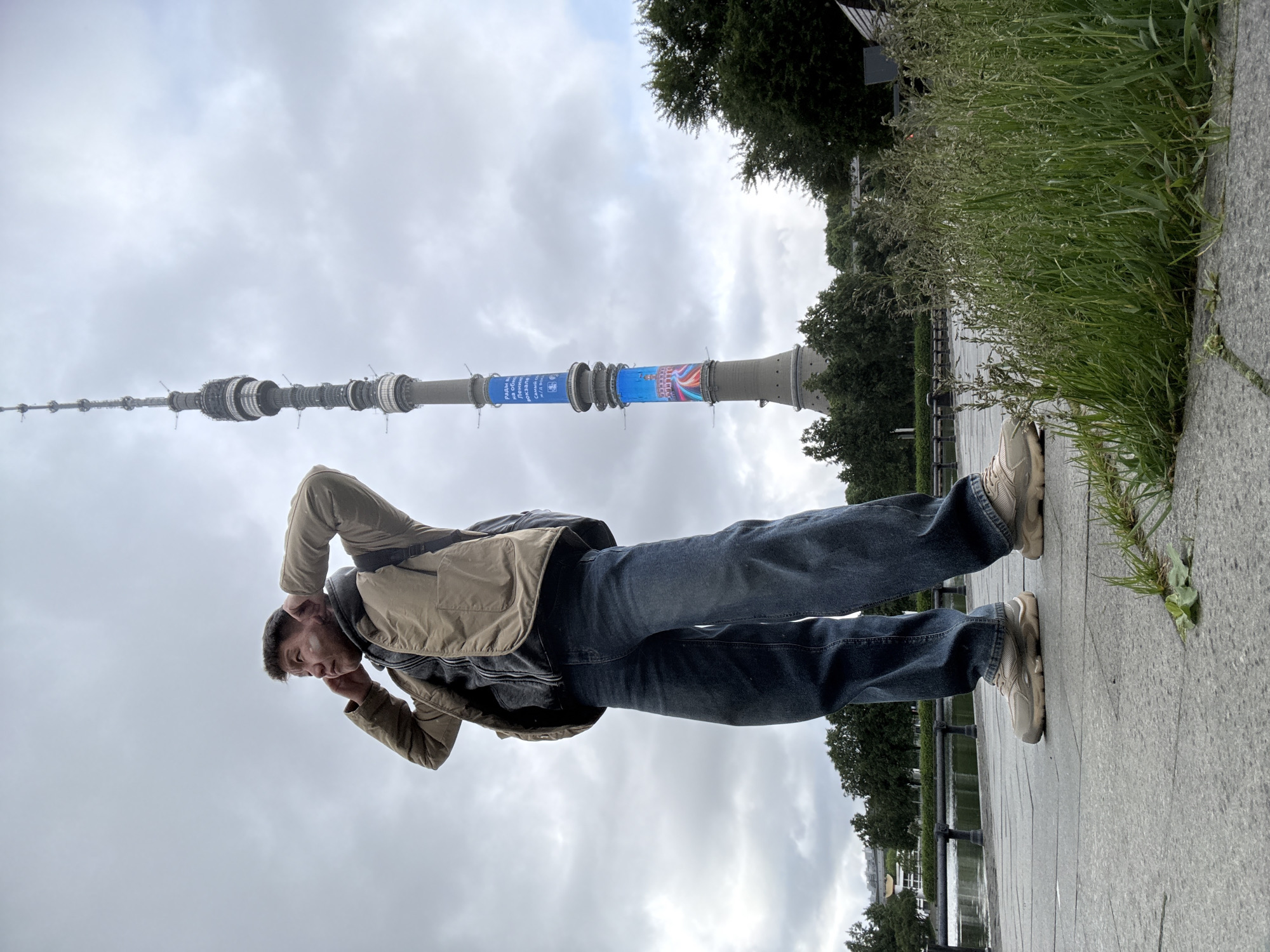 A man in jeans and a beige jacket is standing on a paved path in front of the Ostankino Tower in Khimki, Russia. His hands are raised to his head, and he is looking toward the camera. Behind him, to the right, is some greenery next to a small canal with a black fence. The sky is overcast.