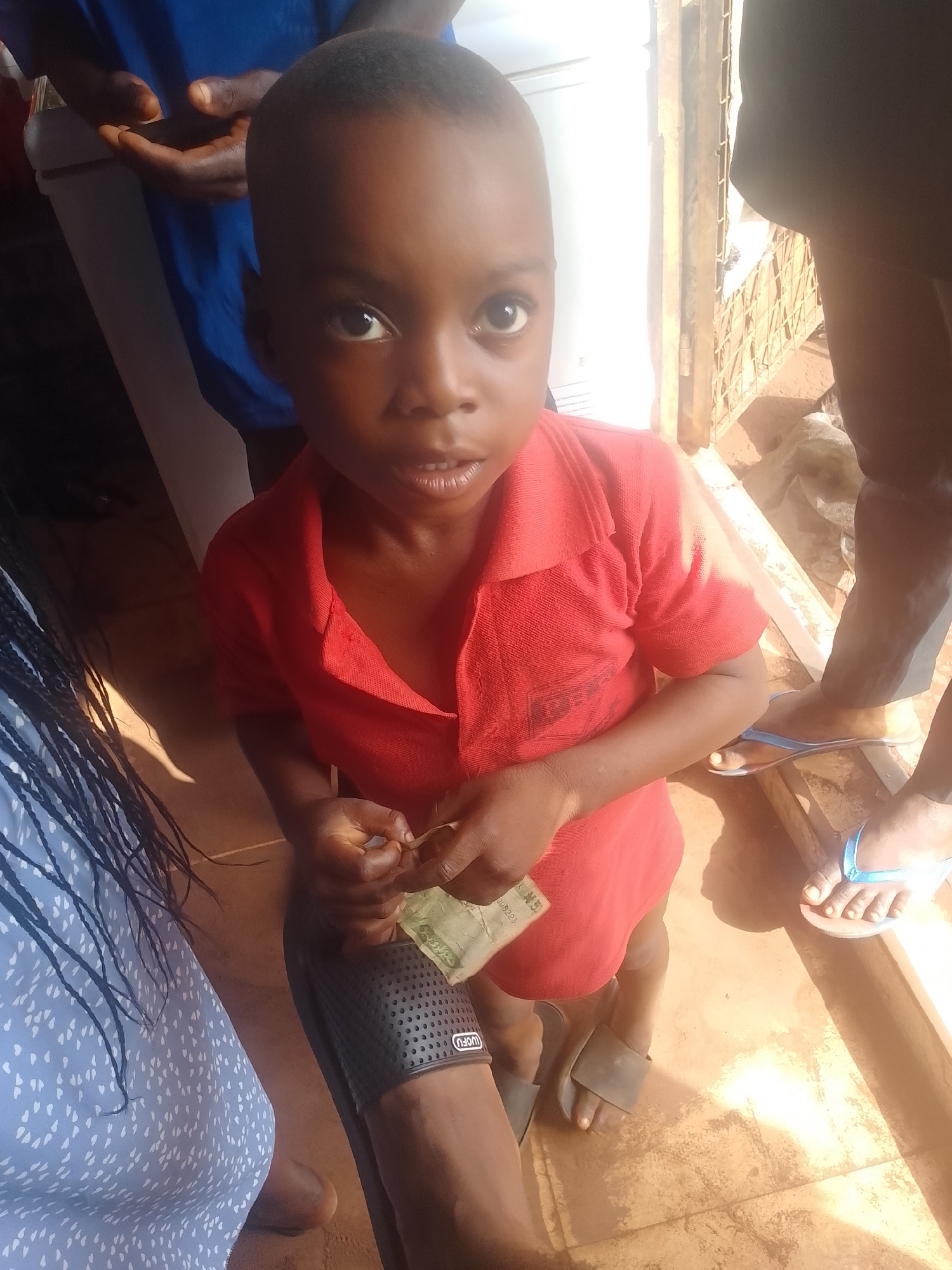 A young, dark-skinned child wearing a red collared shirt stands in what appears to be an outdoor or semi-outdoor setting in Ejule, Nigeria. The child holds what looks like a small amount of Nigerian currency in their hands. The floor is a light brown color. Several other people are partially visible around the child; one person wearing a blue dress with white spots is visible on the left, while another person in dark pants and blue flip-flops is on the right. A man in a blue shirt is partly seen in the upper left part of the image. The background includes a partially obscured building. The light suggests a bright, possibly sunny day.