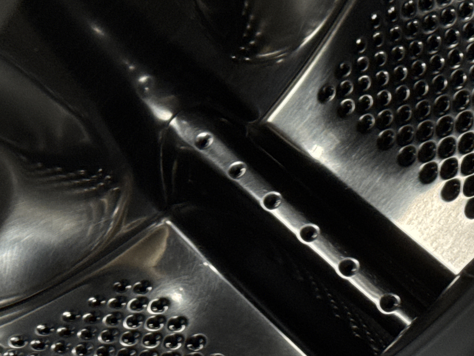 The image is a close-up, abstract view of the interior of a stainless steel washing machine drum. The drum has a patterned surface with many small, raised, rounded dimples, and several rows of small holes are visible along a raised ridge that runs diagonally across the frame. The lighting creates strong reflections and highlights on the metal surfaces, emphasizing the texture and curvature. The overall impression is one of industrial material and design. The image does not depict any recognizable landmarks or features that would allow for its location to be determined within the city of Izhevsk, Russia.