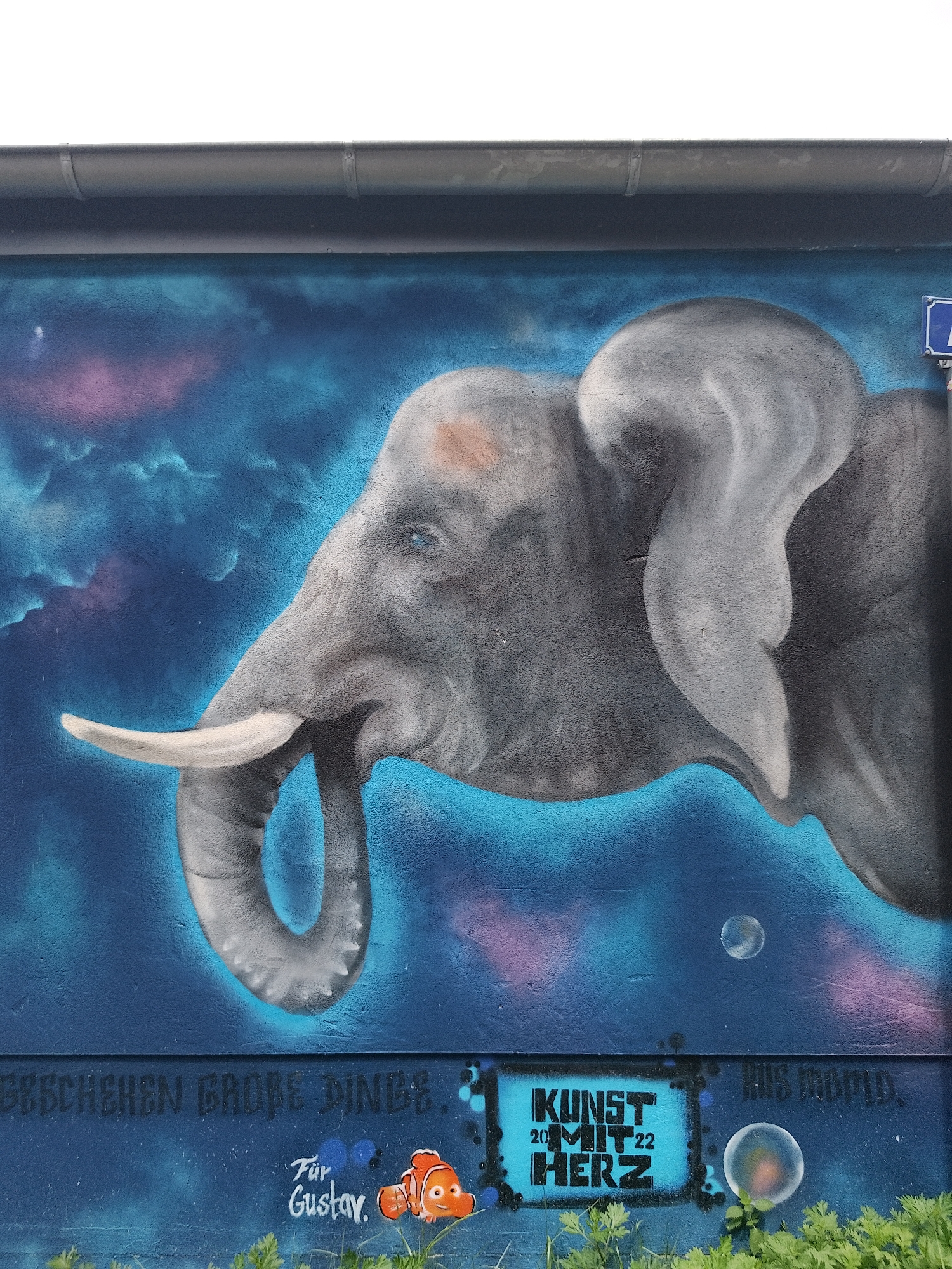 The image captures a mural in an urban environment. An elephant head dominates the scene, painted in shades of gray and blue against a blue background that suggests a cosmic, cloud-like sky. The mural also features details such as bubbles floating around the elephant's head and inscriptions in German. Below the elephant, graffiti-style elements are present. Notably, a small "Finding Nemo" character is depicted, and text reads "Für Gustav." The words "Kunst mit Herz" (Art with Heart) are stenciled in blue. The bottom edge of the mural is partially obscured by greenery. A street sign is visible in the upper right.